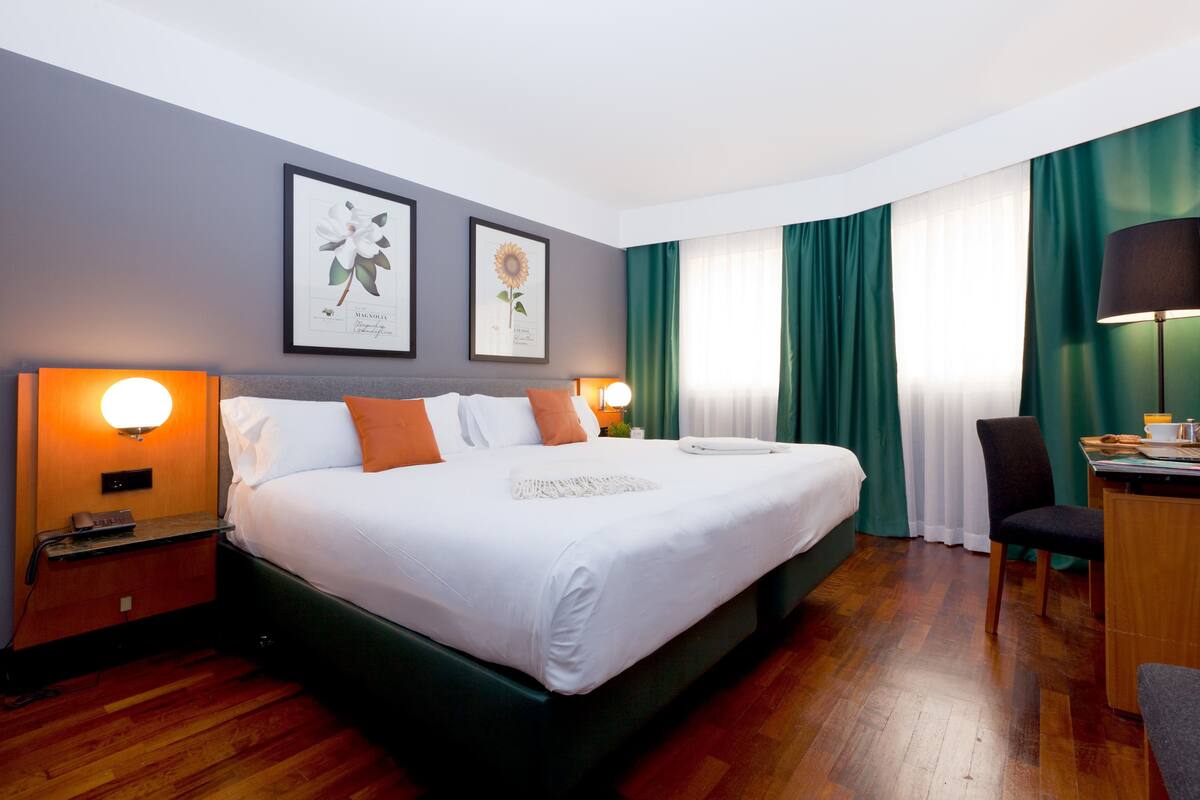 Habitación de hotel con una cama grande, dos mesitas de noche con lámparas, un escritorio con silla y una ventana con cortinas.