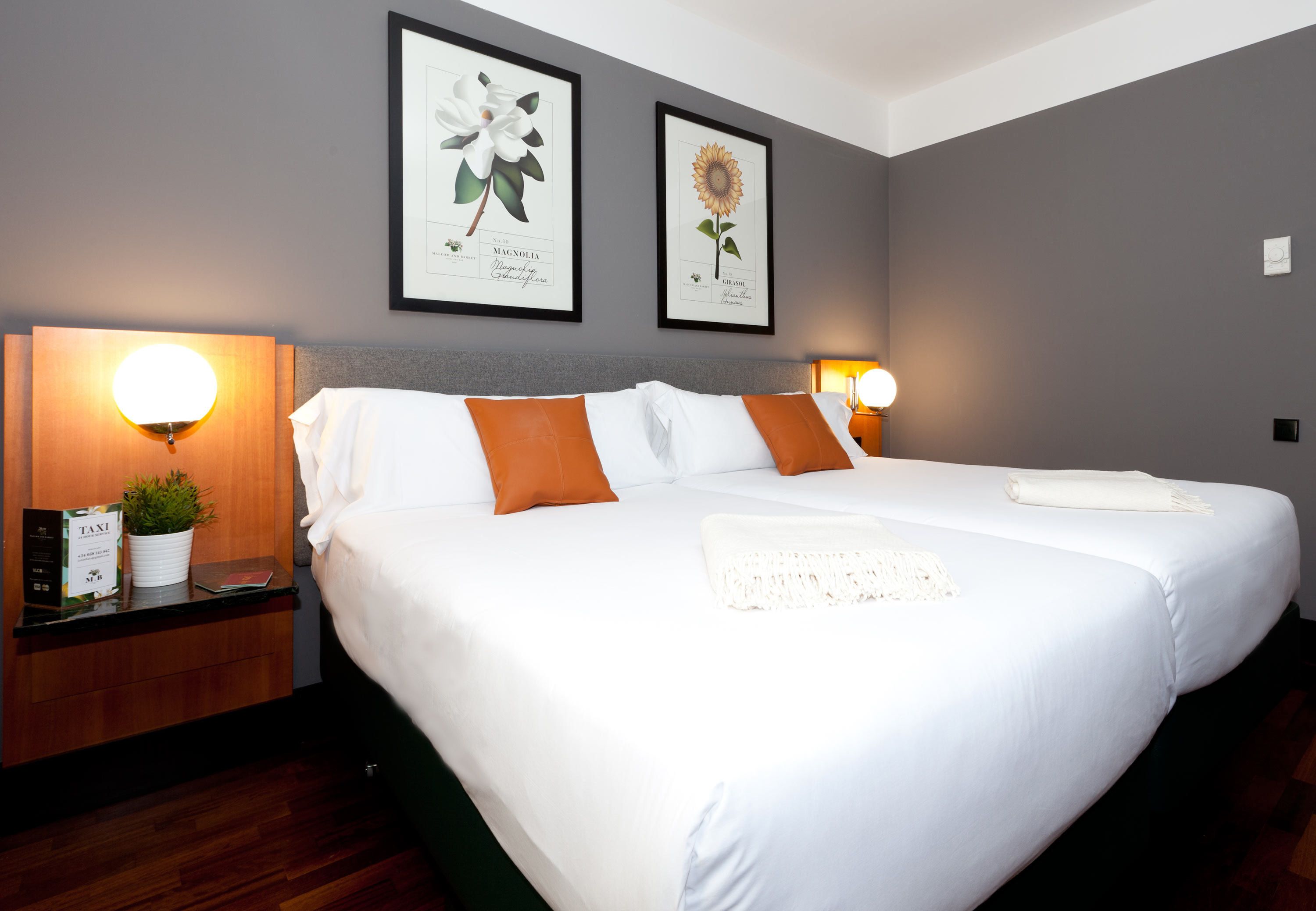 A double bed with white and orange bedding, two framed botanical prints on the wall, and bedside lamps.