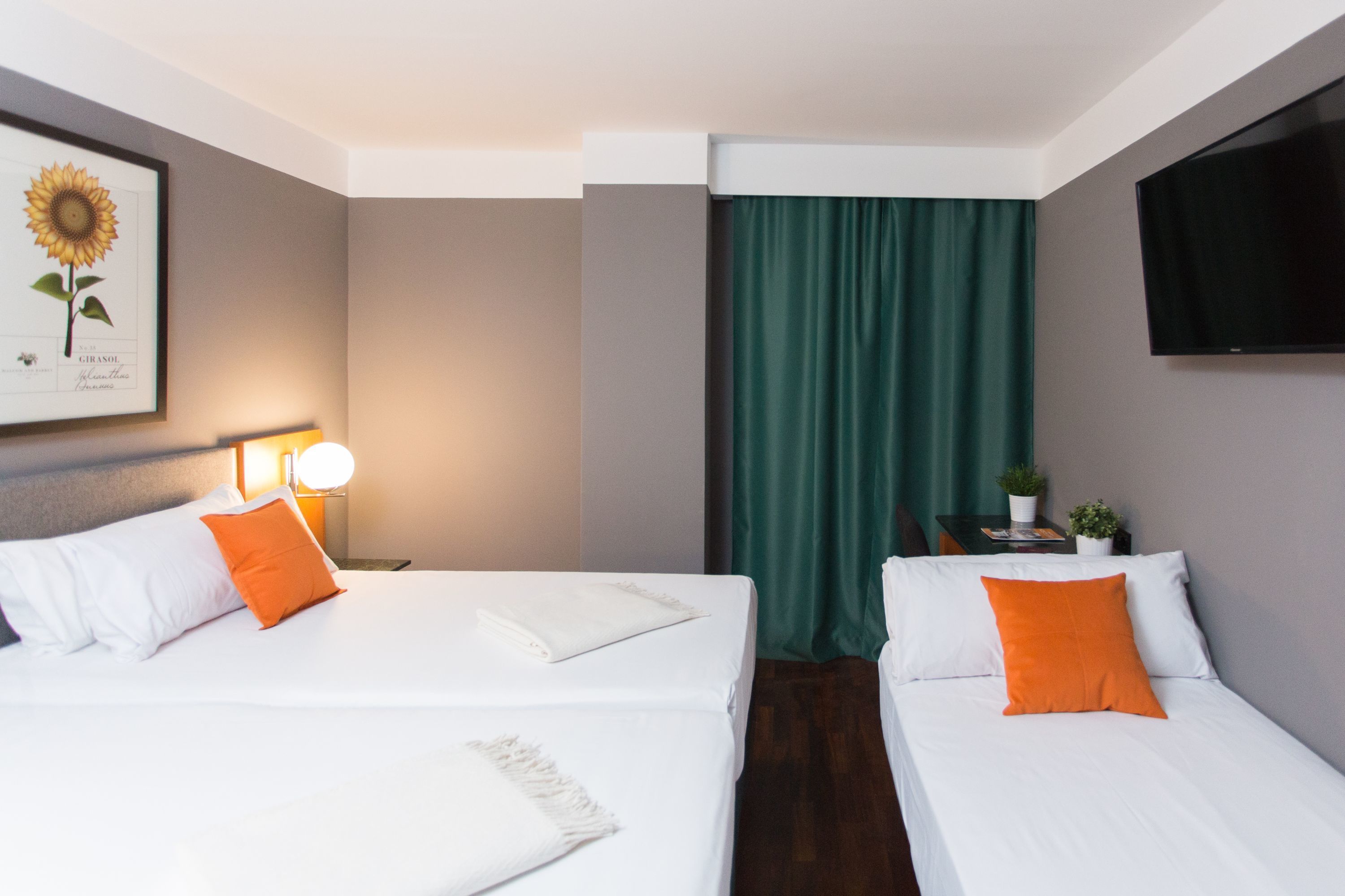 A hotel room with two beds, a flat-screen TV, a framed botanical print, a nightstand with a lamp, and a green curtain.