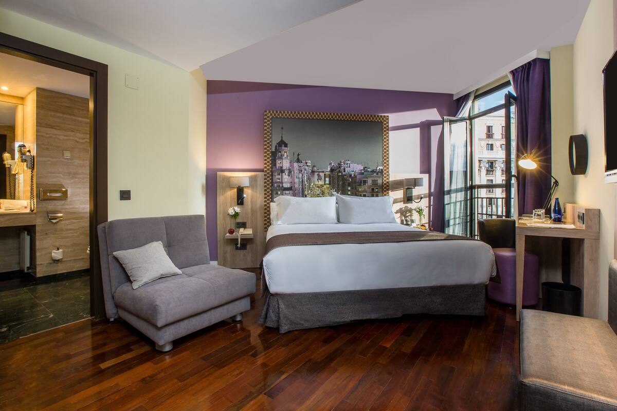 Una habitación de hotel moderna con cama, sofá, escritorio y vistas a un paisaje urbano a través de grandes ventanales.