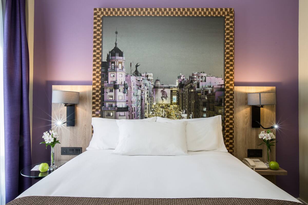 Une chambre d’hôtel avec un grand lit, deux tables de chevet avec des lampes, un tableau représentant un paysage urbain accroché au mur et des murs de couleur violette.