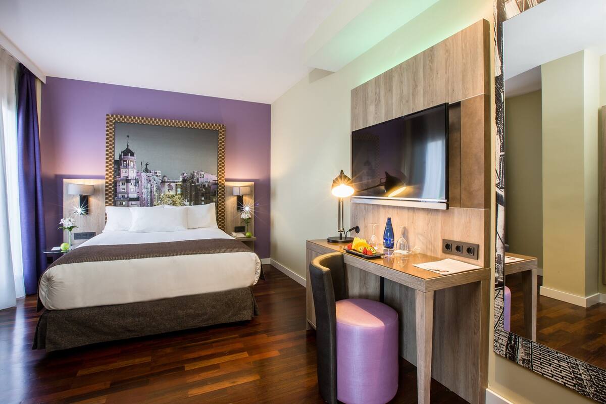 Habitación de hotel con cama, escritorio, silla, espejo y televisor.