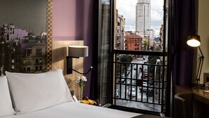 Hotelkamer met een bed, nachtkastje, lamp, vaas met bloemen en raam met uitzicht over de stad.