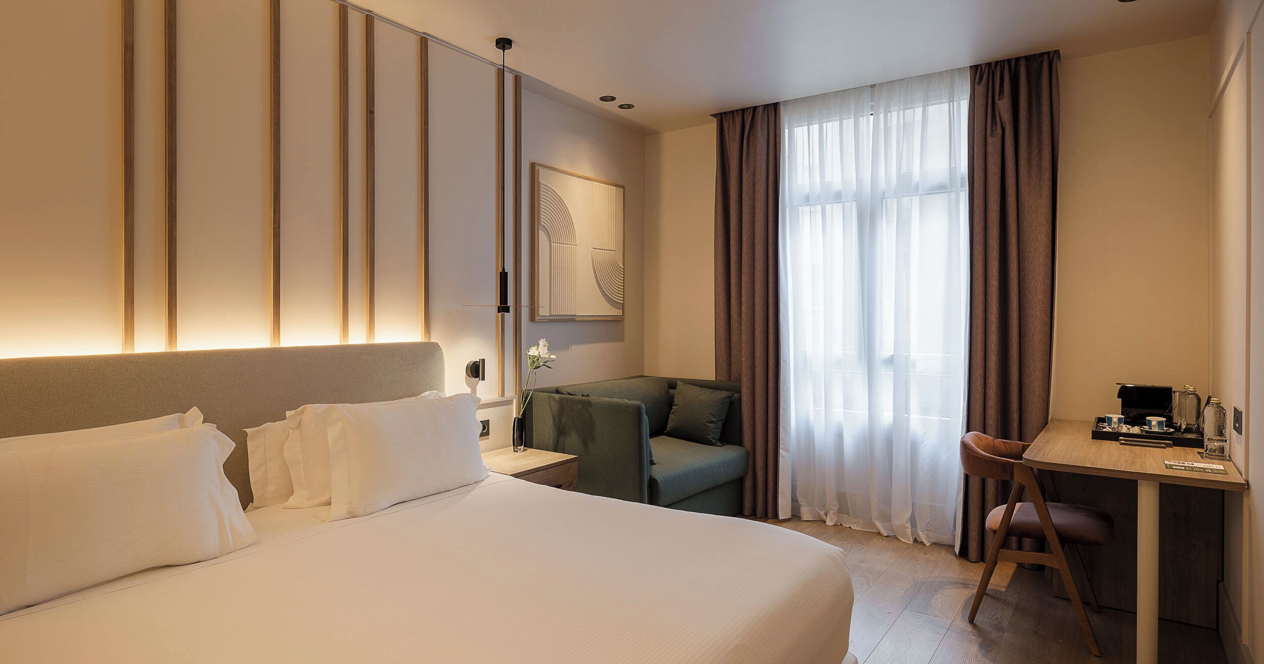 Habitación de hotel con una cama grande, un escritorio, una silla, un sofá y un ventanal con cortinas.