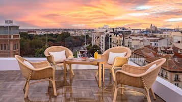 Un balcón con mobiliario de mimbre y una mesa, con vista a un paisaje urbano al atardecer.