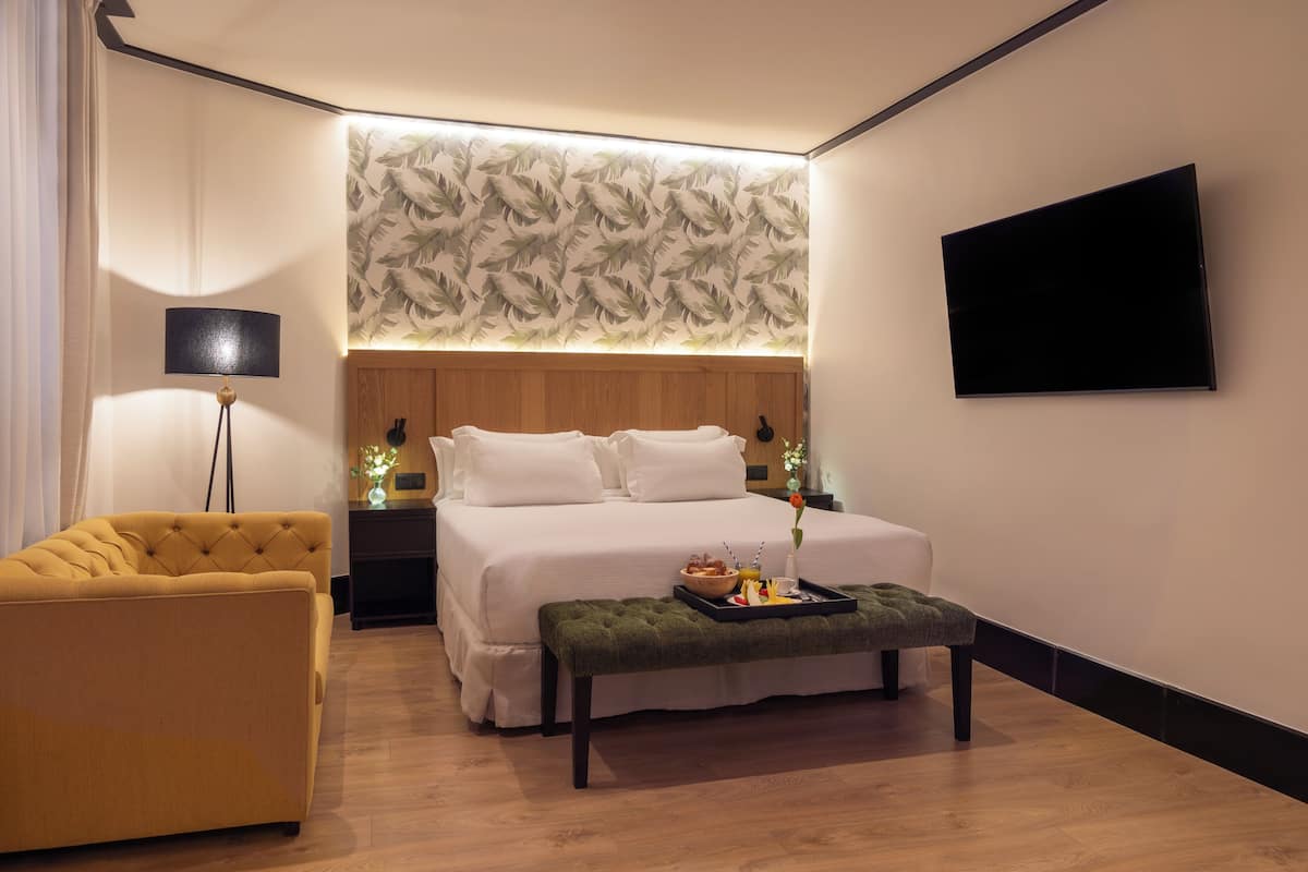 A modern hotel room with a large bed, a sofa, a flat-screen TV, and a wall-mounted lamp.