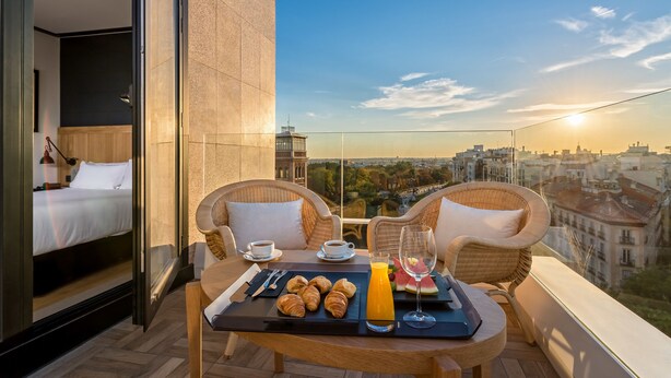 A balcony with a breakfast setup including croissants, juice, and a glass of water, overlooking a cityscape at sunset. - H10 Puerta de Alcalá 4* Sup (Madrid)