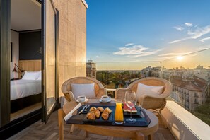 Un balcón con desayunos que incluyen cruasanes, jugo y un vaso de agua, con vistas a un paisaje urbano al atardecer.