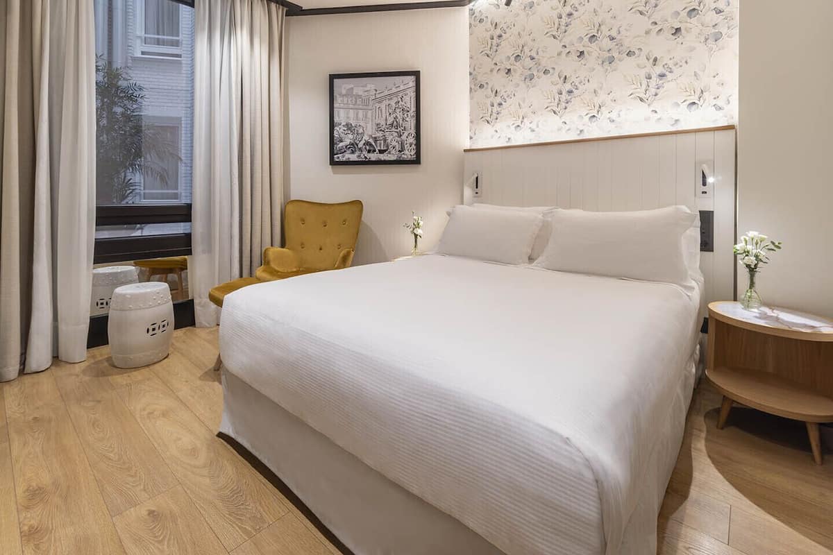 A hotel room with a large bed, a chair, a small table, and a window with curtains.