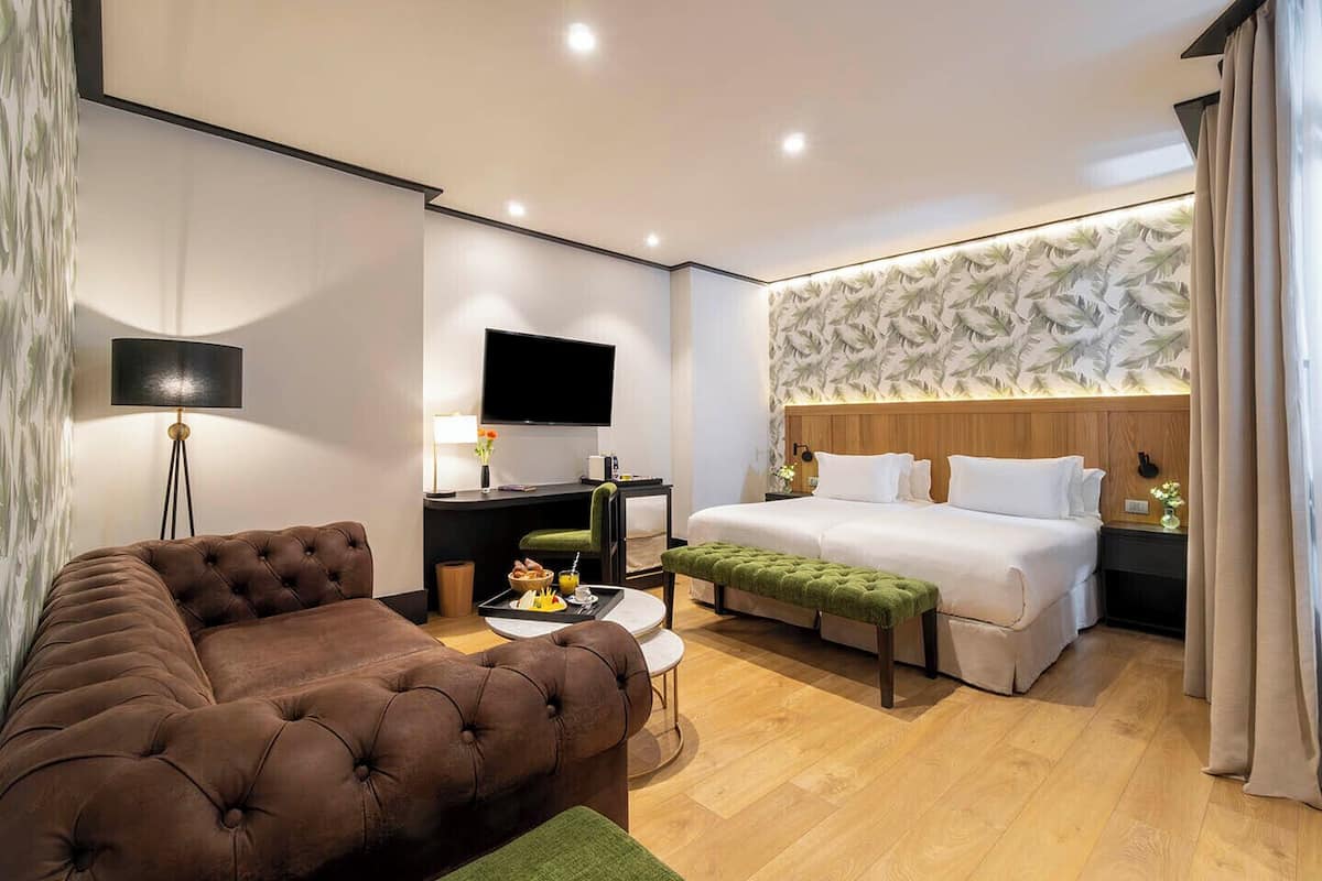A hotel room with a large bed, a sofa, a desk with a flat-screen TV, and a wall-mounted TV.