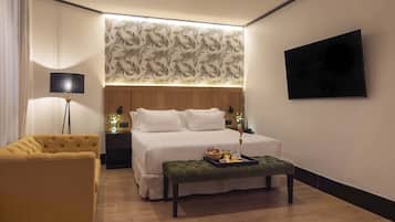 Een hotelkamer met een bed, een nachtkastje met lamp, een aan de muur bevestigde televisie, een gele fauteuil, en een dienblad met snacks op de vloer.