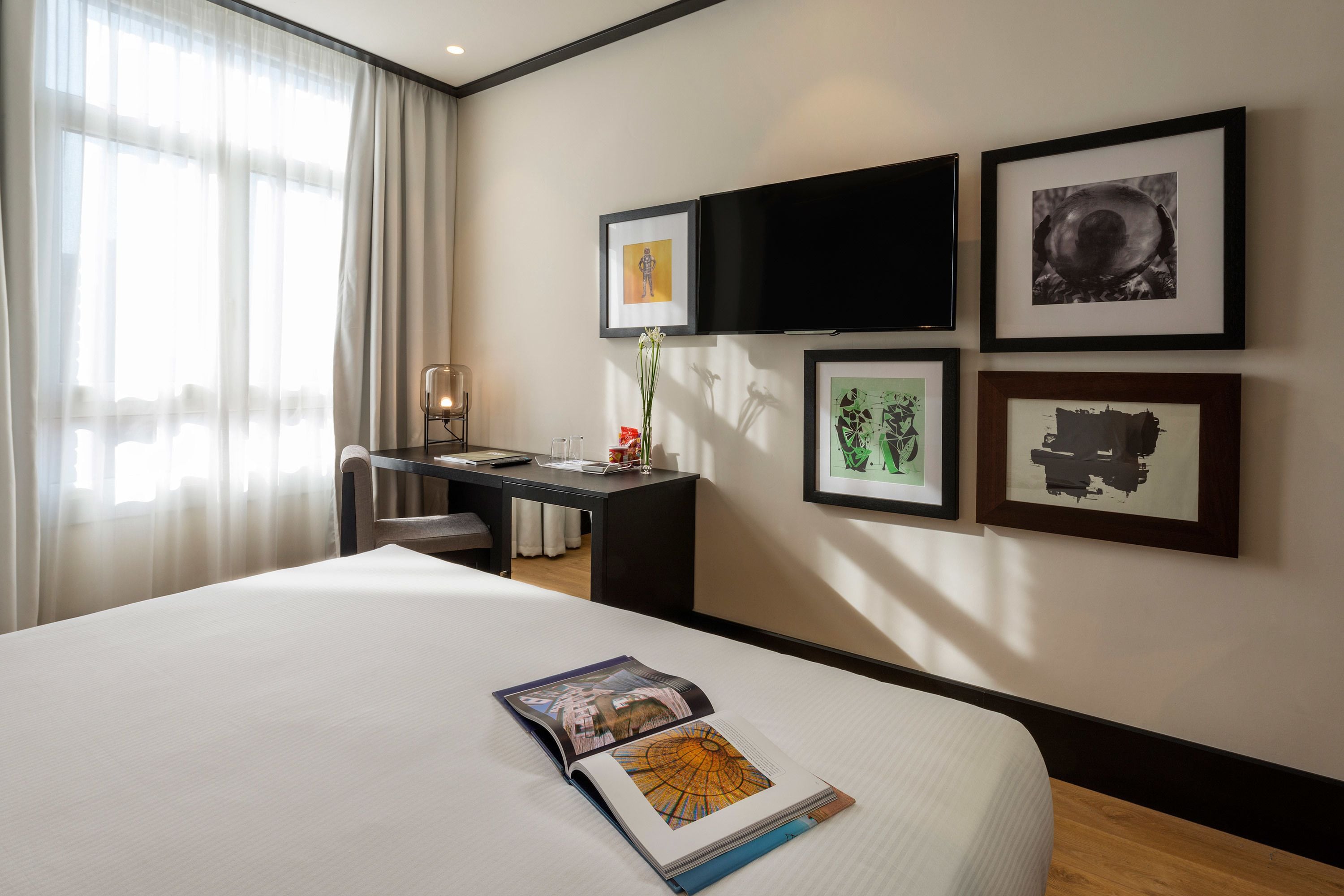 Habitación de hotel con una cama grande, un escritorio con silla, un televisor de pantalla plana en la pared y varias fotografías enmarcadas.