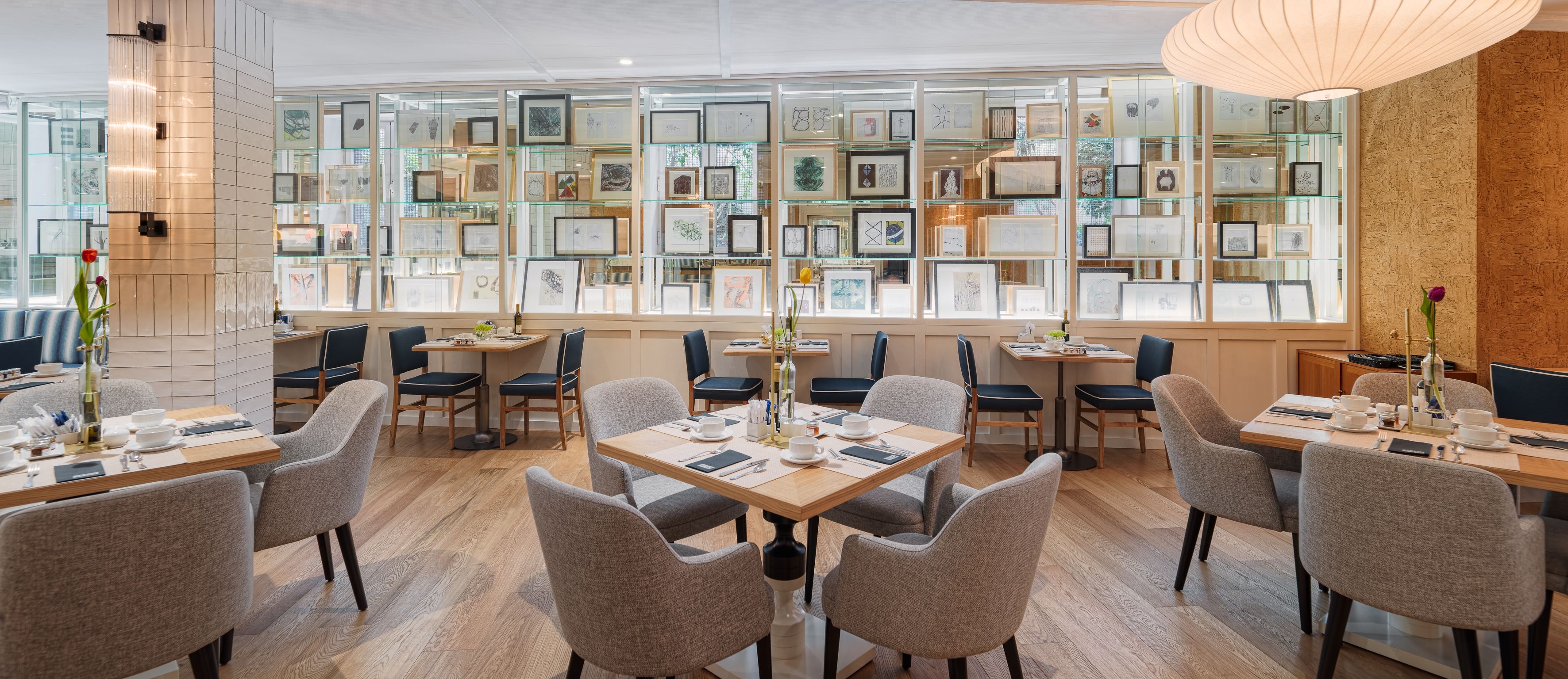 Un comedor moderno con varias mesas y sillas, una pared de vidrio con obras de arte enmarcadas y piso de madera.
