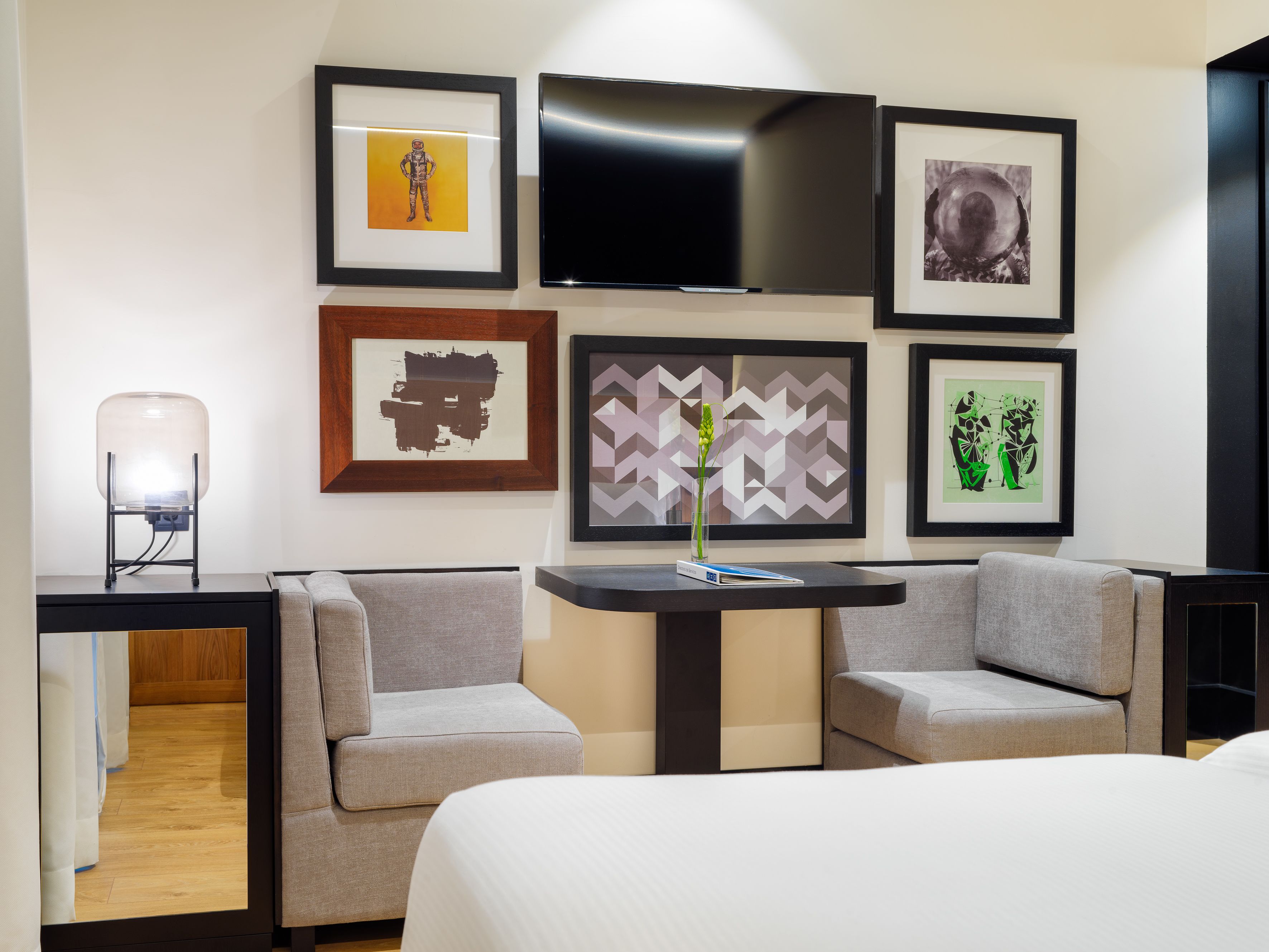 Habitación de hotel con cama, dos sillones, una mesita, televisor y varias obras de arte enmarcadas en la pared.