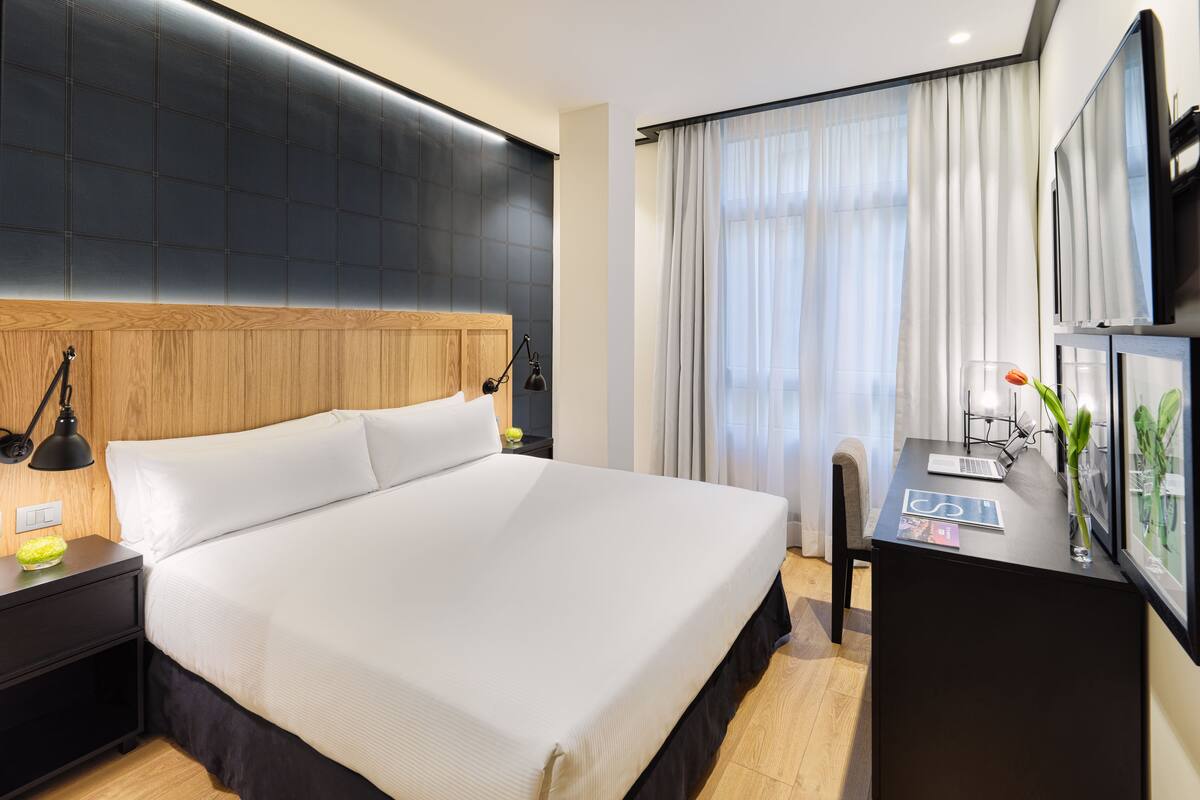 A hotel room with a large bed, a desk with a laptop, a nightstand with a lamp, and a window with curtains.