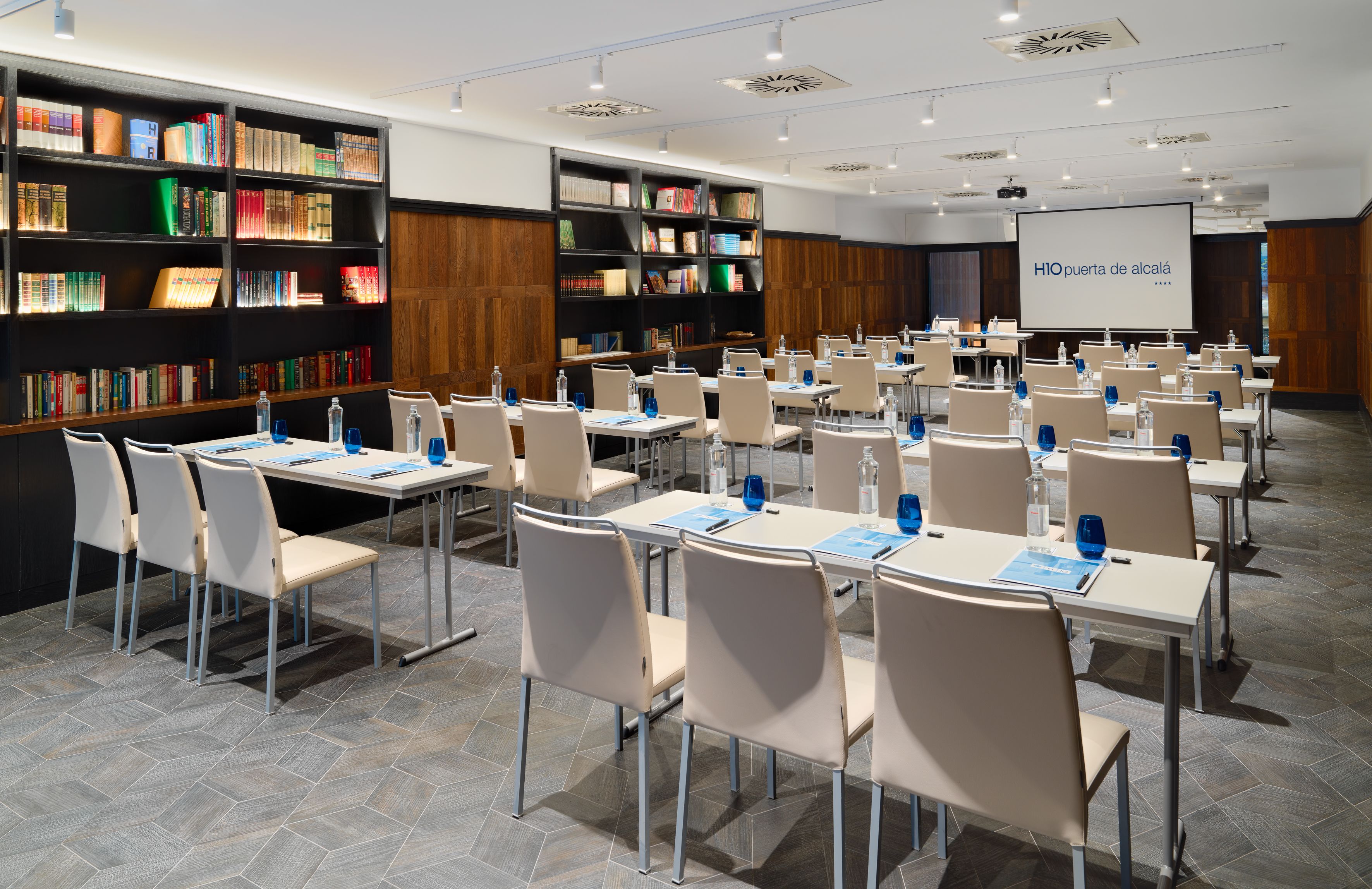 Un salón de conferencias con filas de mesas y sillas, una pantalla de proyector y estanterías llenas de libros.