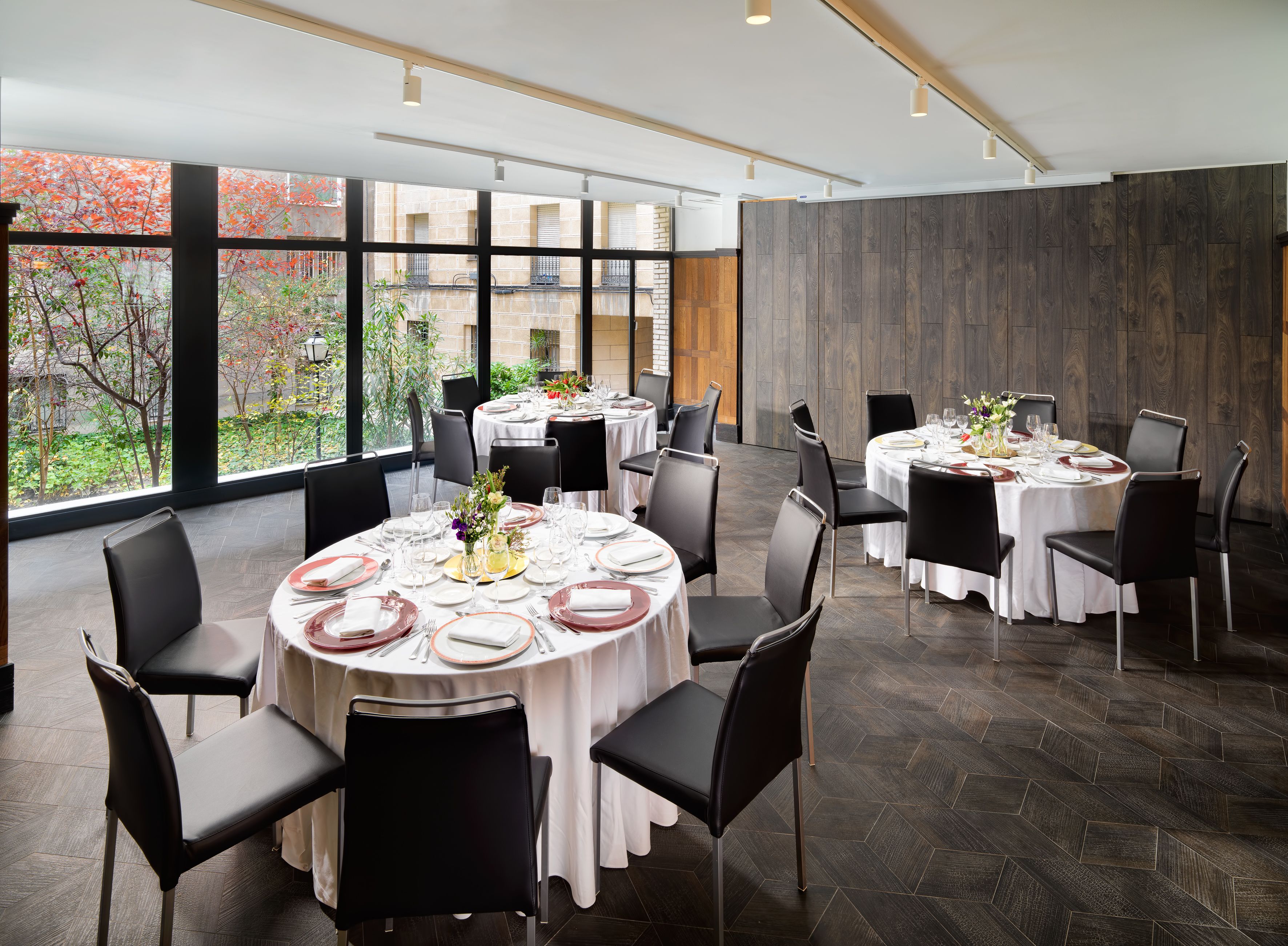 Een restaurant met ronde tafels klaar voor diners, grote ramen met uitzicht op bomen, en een modern interieur.