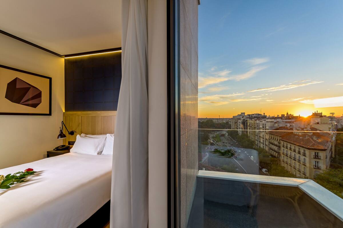 A hotel room with a bed, a bedside table, a lamp, a framed picture, and a window with a city view at sunset.