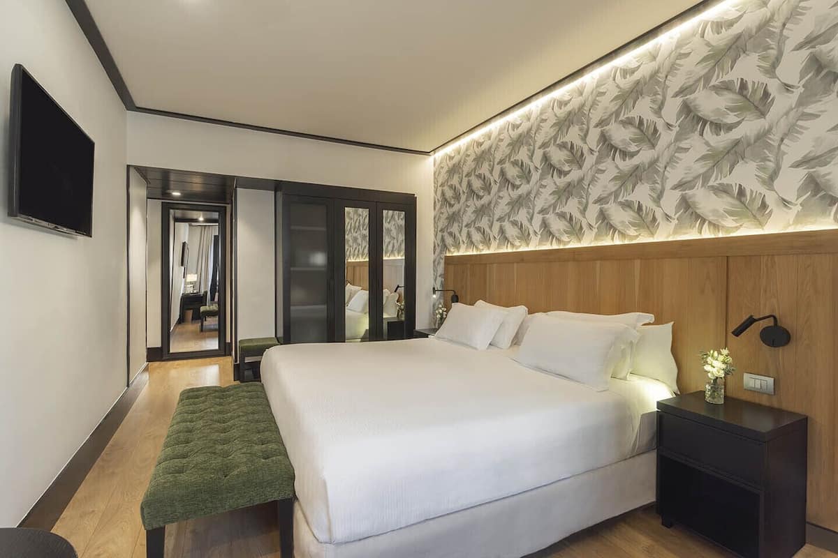 A modern hotel room with a large bed, a green bench, a wooden headboard, a nightstand, and a wall-mounted TV.