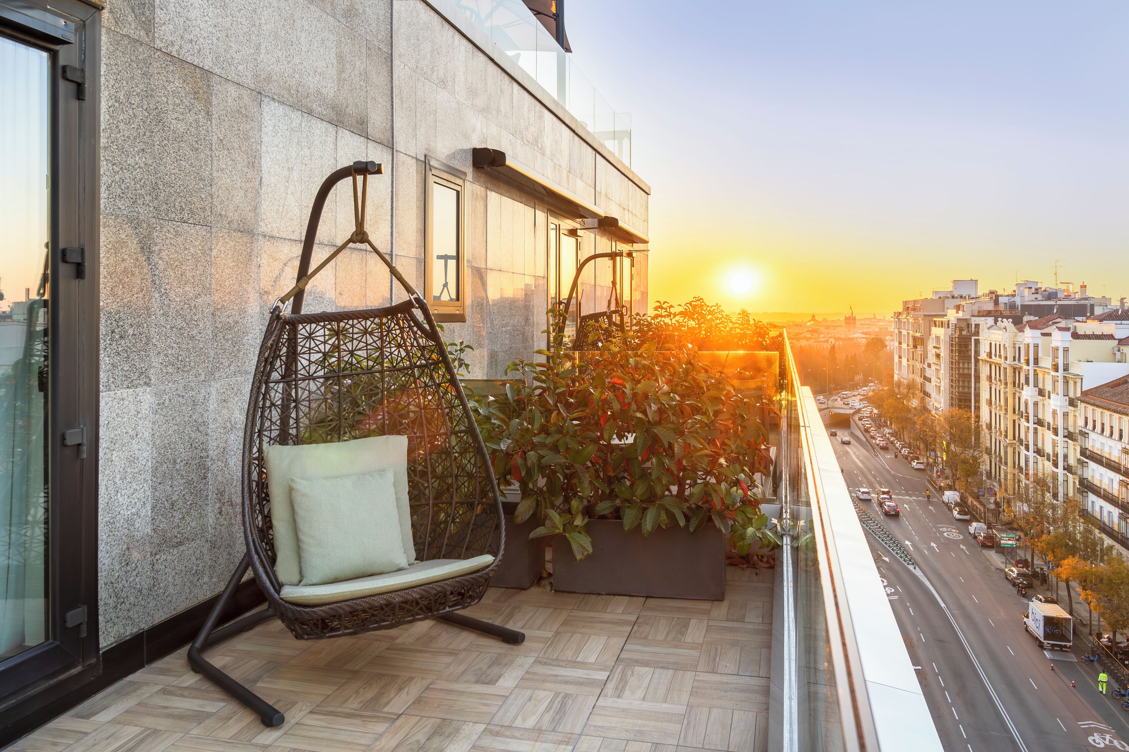 A balcony with a hanging chair, potted plants, and a city view at sunset.