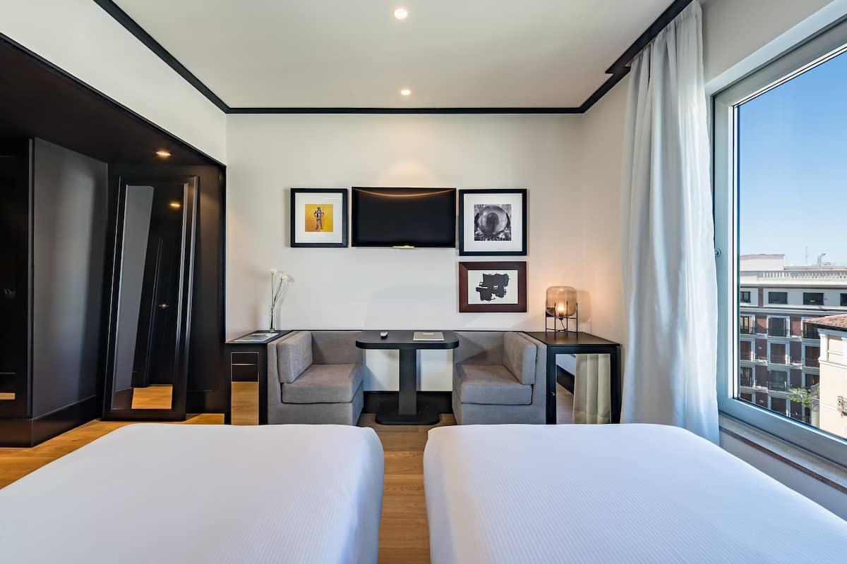 A modern hotel room with two beds, a desk with two chairs, a TV, and framed artwork on the wall.