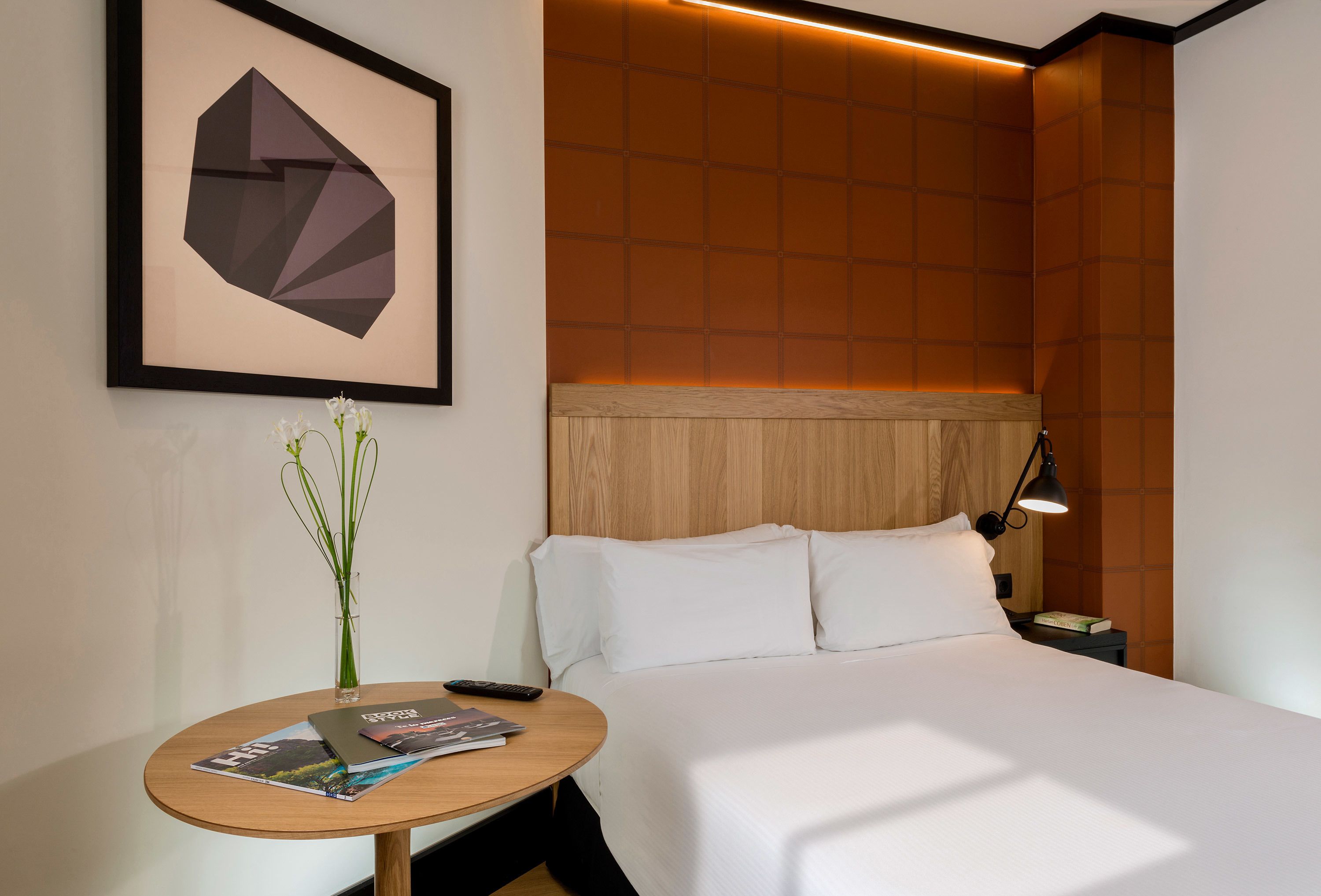 Habitación de hotel con cama, mesita de noche con lámpara, una obra de arte abstracta enmarcada y un jarrón con flores.