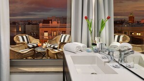 A hotel room with a balcony, wicker furniture, a white sink, and a view of the cityscape at sunset. - H10 Puerta de Alcalá 4* Sup (Madrid)