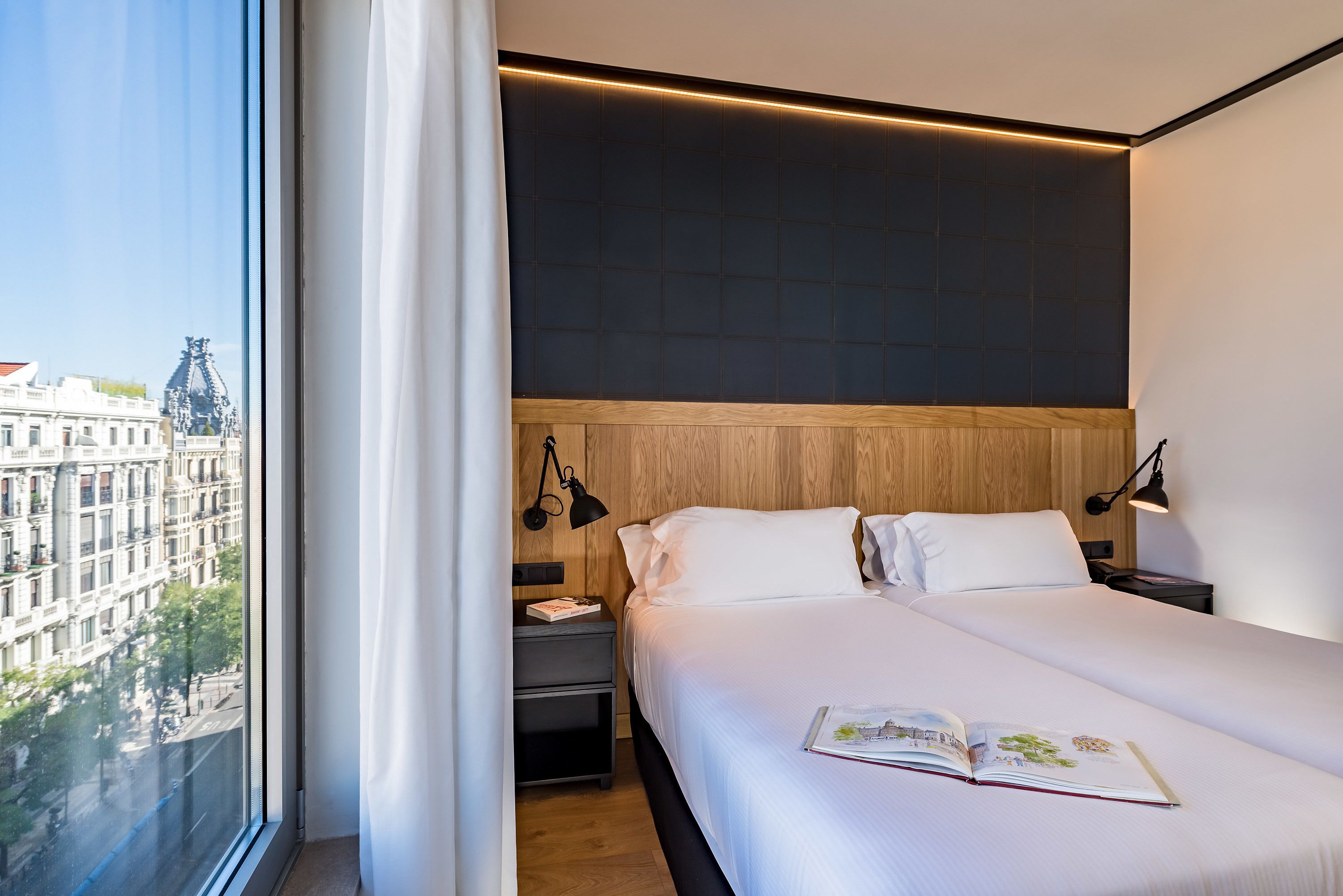 Ein modernes Hotelzimmer mit großem Fenster, einem ordentlich bezogenen Bett, einem Nachttisch und einem Holz-Kopfteil.
