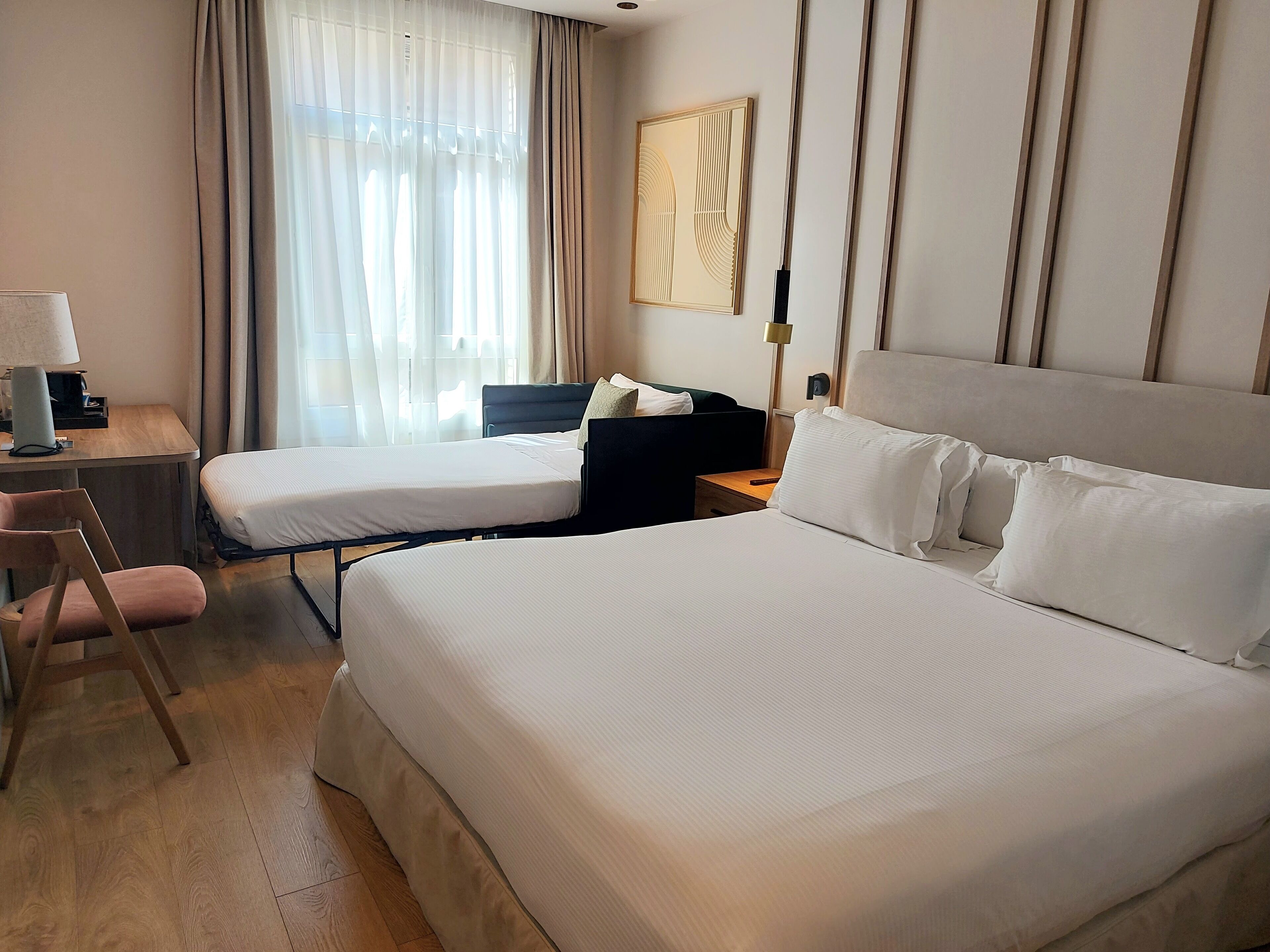 Habitación de hotel con dos camas, un escritorio con lámpara, una silla y una ventana con cortinas.