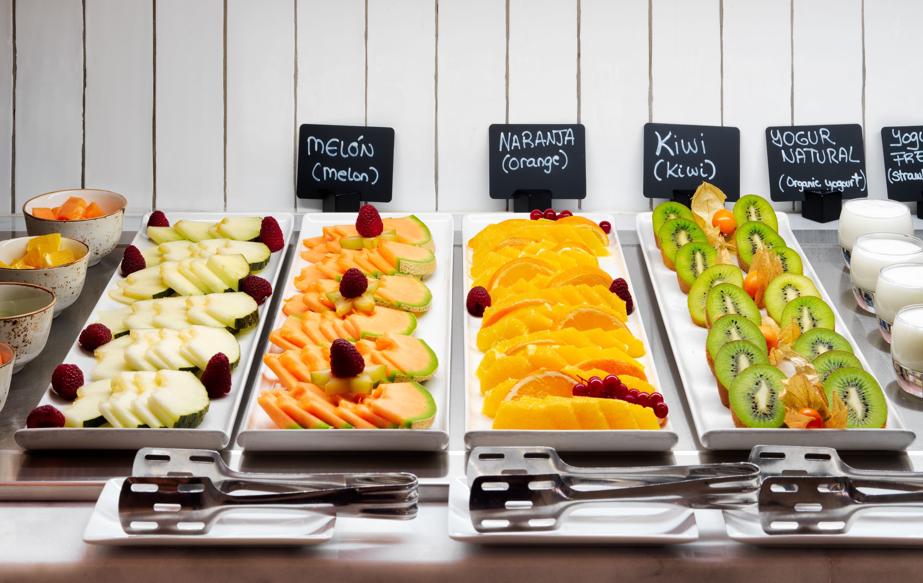 A buffet with various fruit platters labeled with names like melon, orange, kiwi, and yogurt.