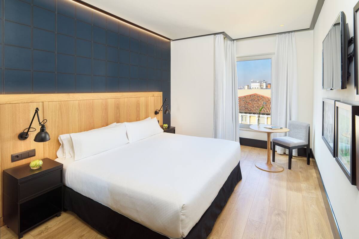 A hotel room with a large bed, a bedside table, a small table with a chair, a window with curtains, and a TV mounted on the wall.
