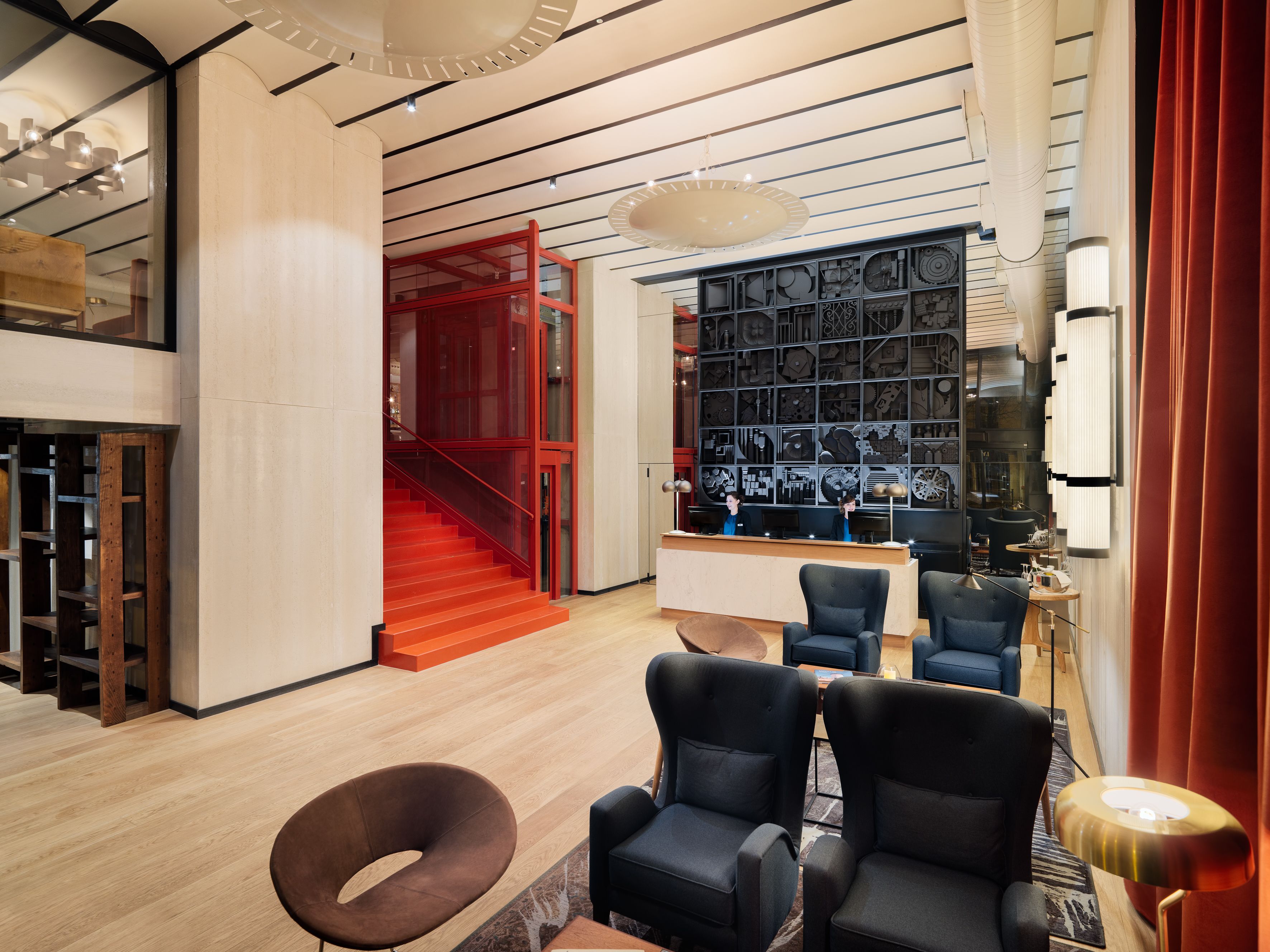 A modern hotel lobby with a wooden floor, a red staircase, and a reception desk with two staff members.