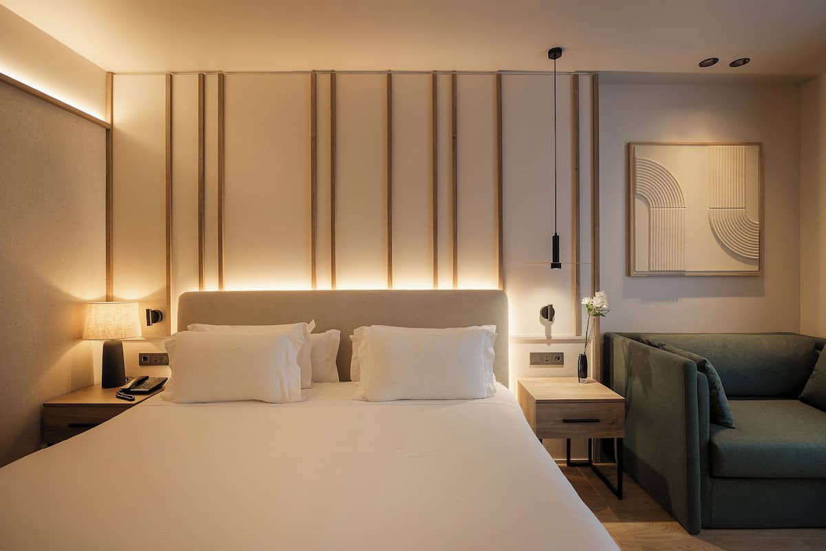 A modern hotel room with a large bed, bedside tables, a desk, and a green armchair.