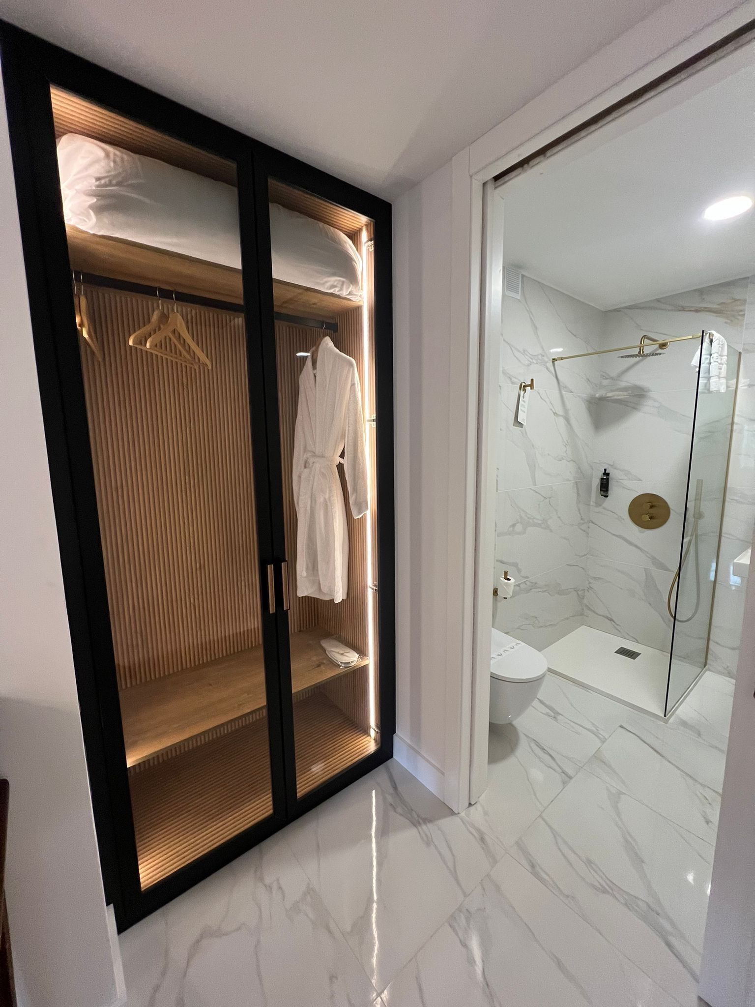 Un baño moderno con ducha envuelta en vidrio, inodoro blanco y tocador de madera con una bata colgada.