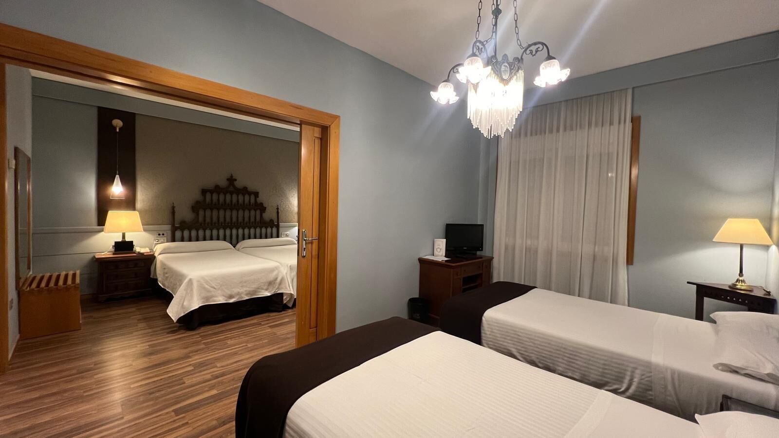 A hotel room with two beds, a wooden floor, a chandelier, a television, and a mirror.