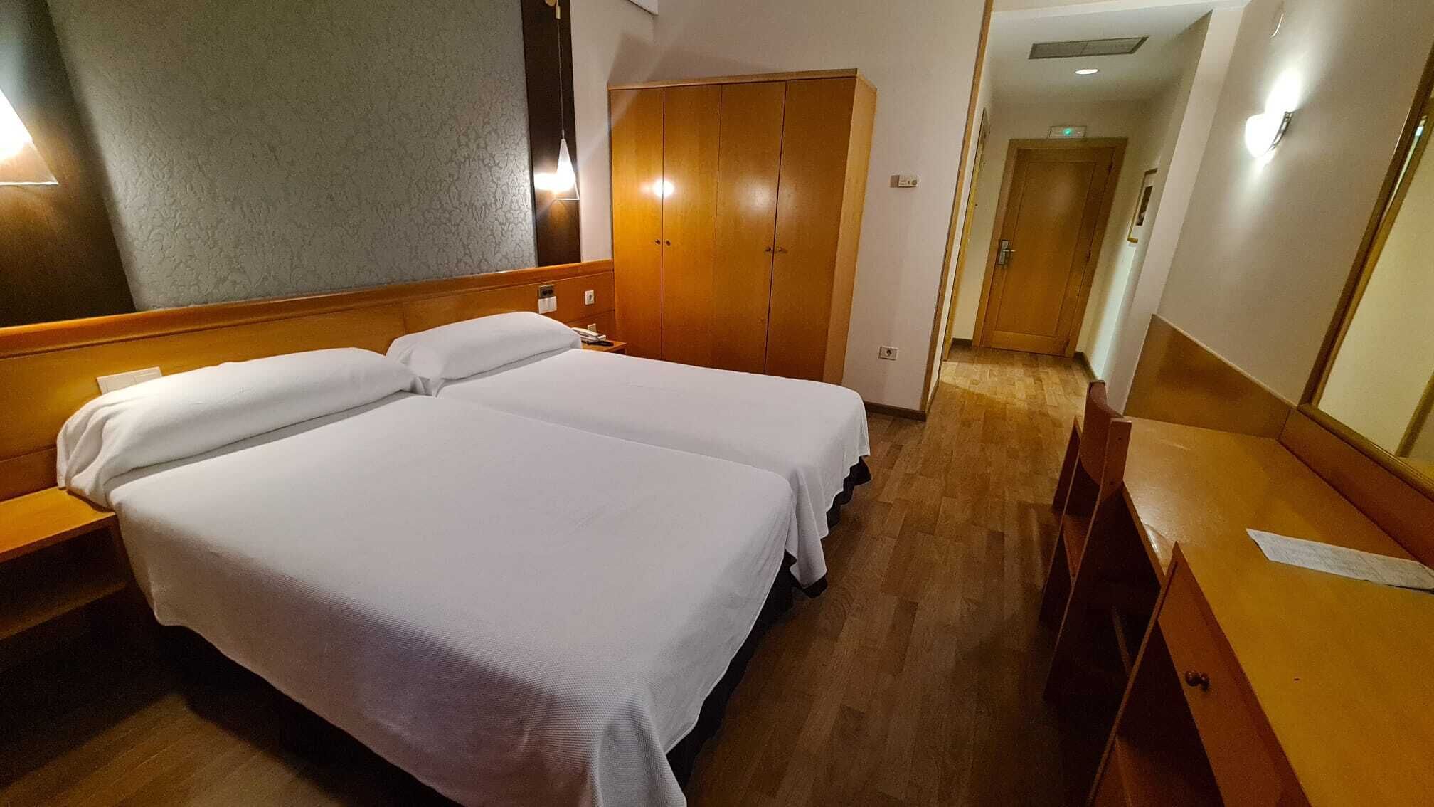 A hotel room with two beds, a wooden wardrobe, a desk with a chair, and a mirror.