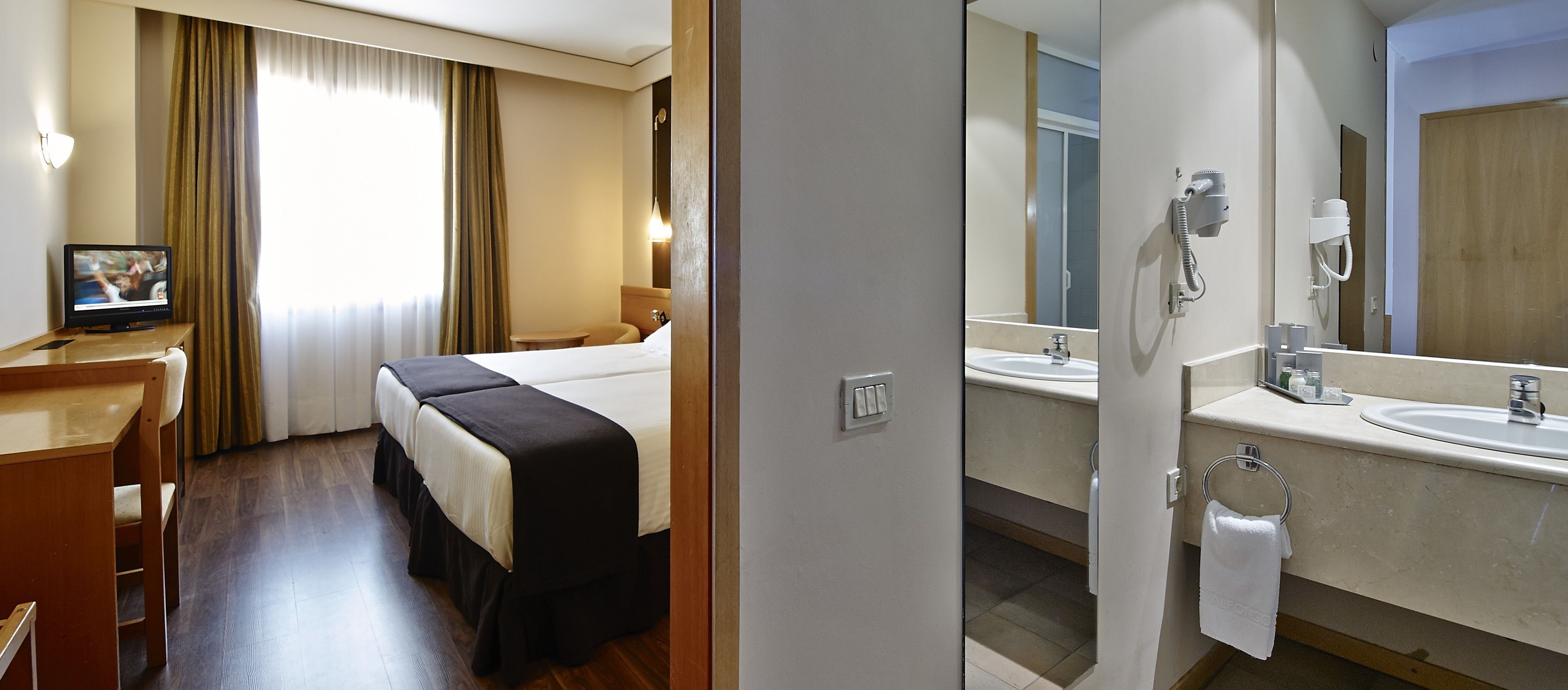 Una habitación de hotel con cama, escritorio, televisor, baño con lavamanos y espejo, y ducha.