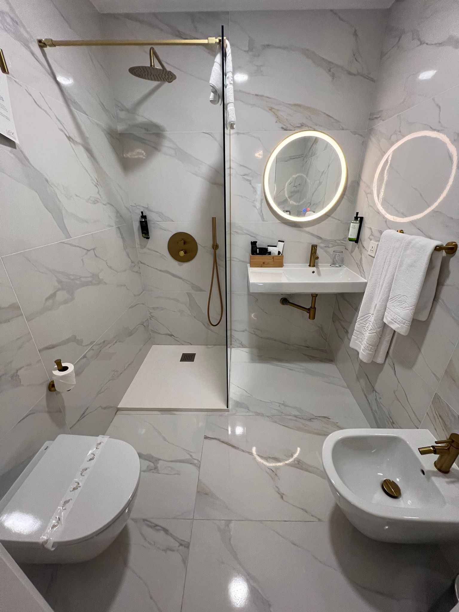 Ein modernes Badezimmer mit einer einteiligen Duschkabine, Toilette, Bidet und zwei Waschbecken mit goldfarbenen Armaturen.