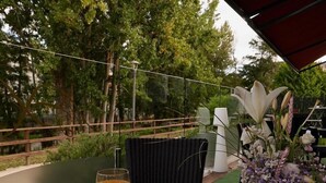Una mesa dispuesta con un croissant, café, jugo de naranja y un vaso de jugo de naranja, con un florero de flores y vistas a árboles y un camino.