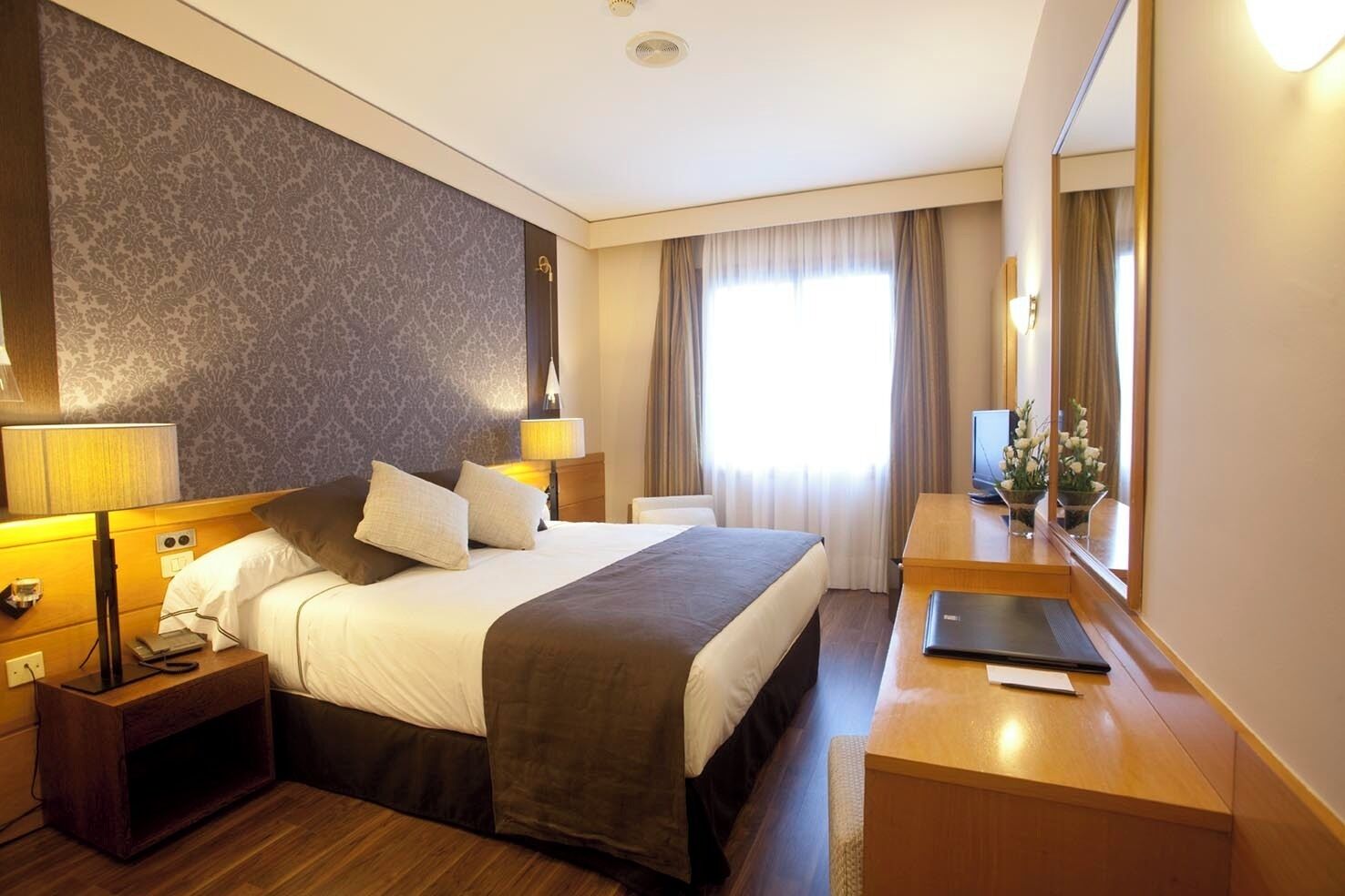 Hotelkamer met een groot bed, bedlampen, een bureau met een laptop, een spiegel en een raam met gordijnen.