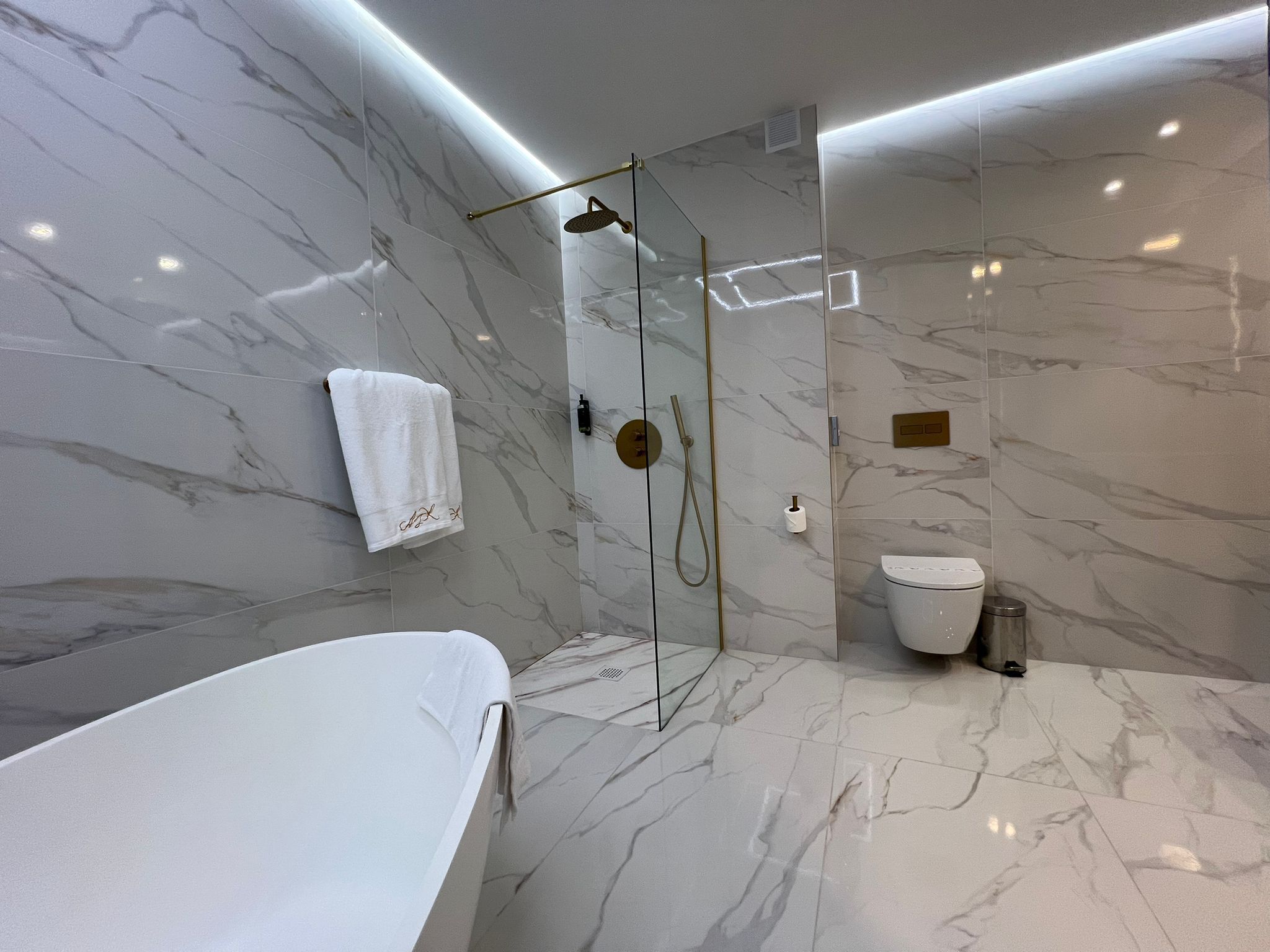 A modern bathroom with a marble floor and walls, a glass-enclosed shower, a white bathtub, and a toilet.