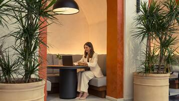 Une femme est assise à une table, un ordinateur portable devant elle, et elle est entourée de plantes en pot. L’ensemble se trouve dans un intérieur moderne, avec un canapé et une lampe.