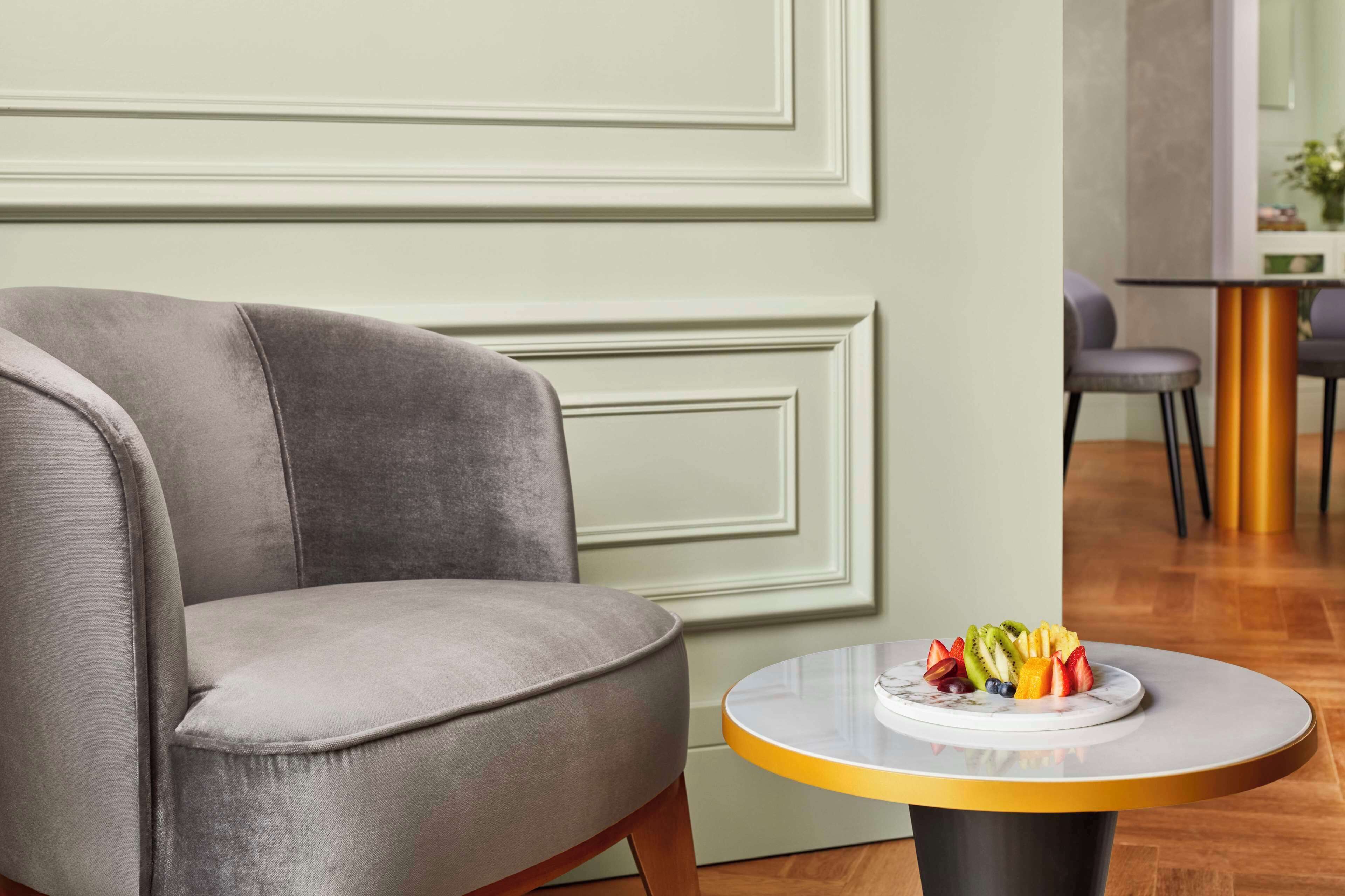 Un salon moderne avec un fauteuil gris, une table ronde sur laquelle est disposé un plateau de fruits, et un coin repas à l’arrière-plan.