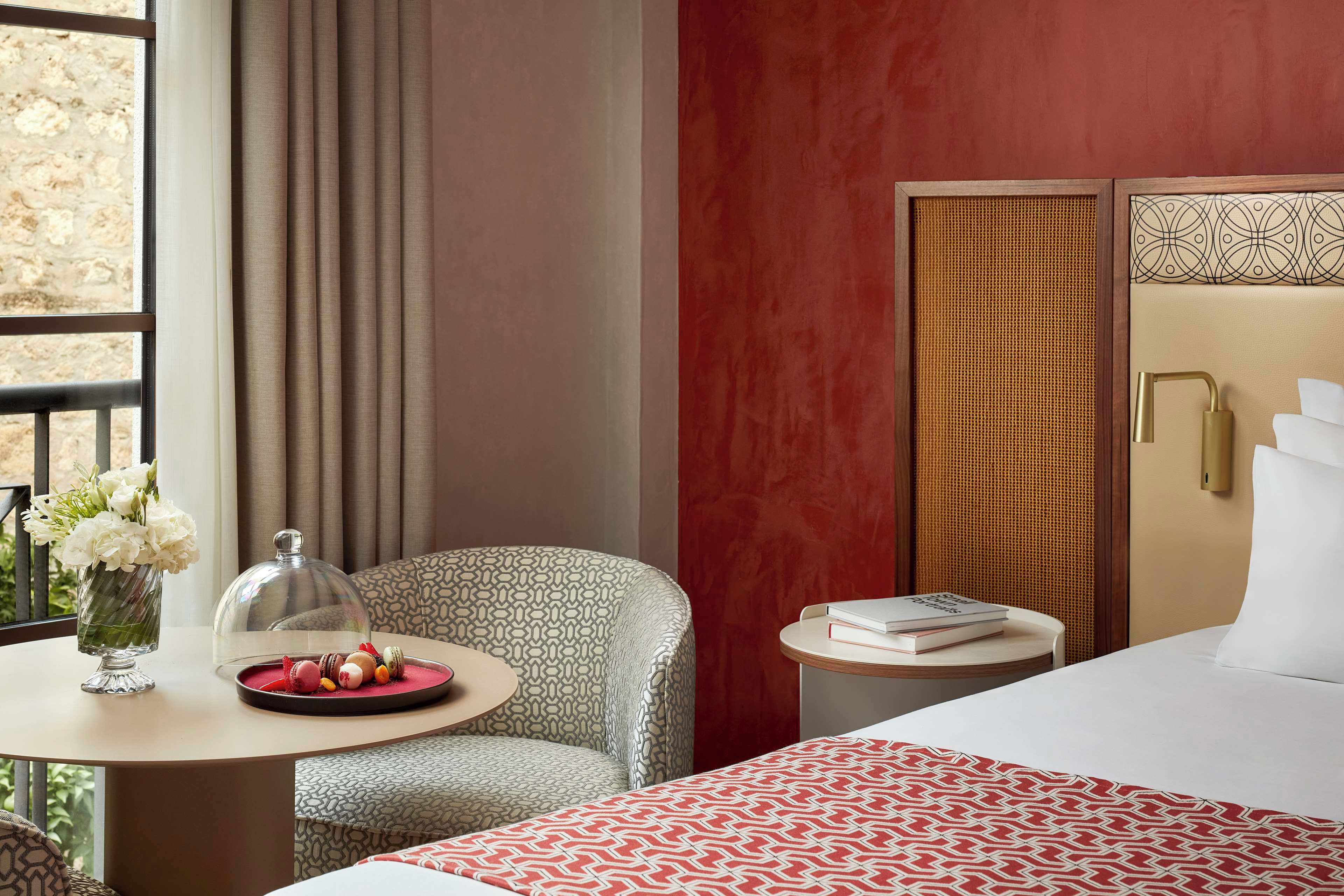 Una habitación de hotel con una cama, una mesa redonda con una bandeja de pasteles, una silla y un jarrón de flores.