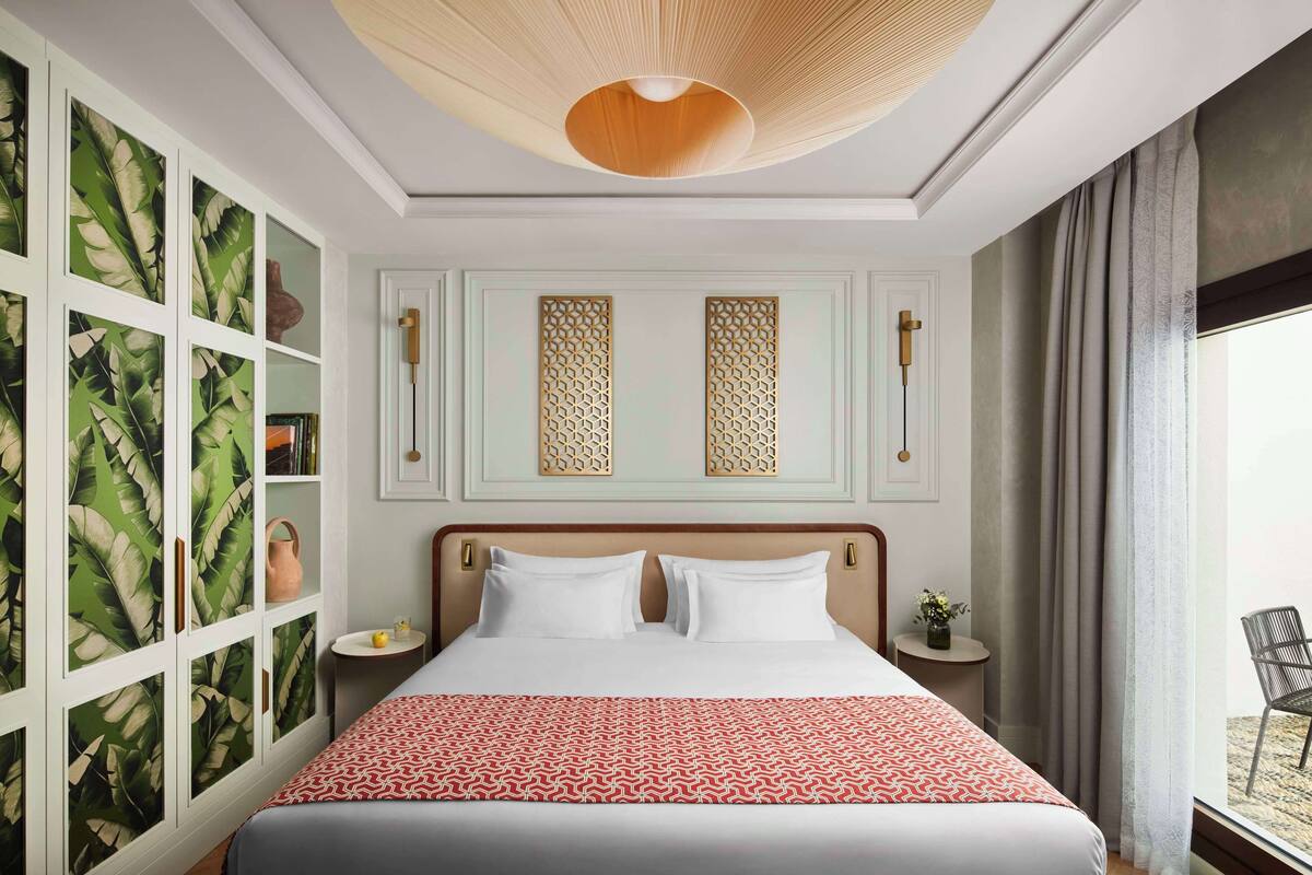 Una habitación de hotel con una cama, una mesita de noche, una pequeña mesa con un florero y un panel decorativo en la pared, además de una ventana con cortinas.