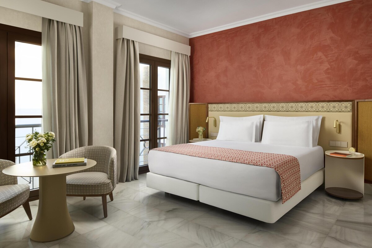 A hotel room with a large bed, a round table, a chair, and a view of the city through the window.
