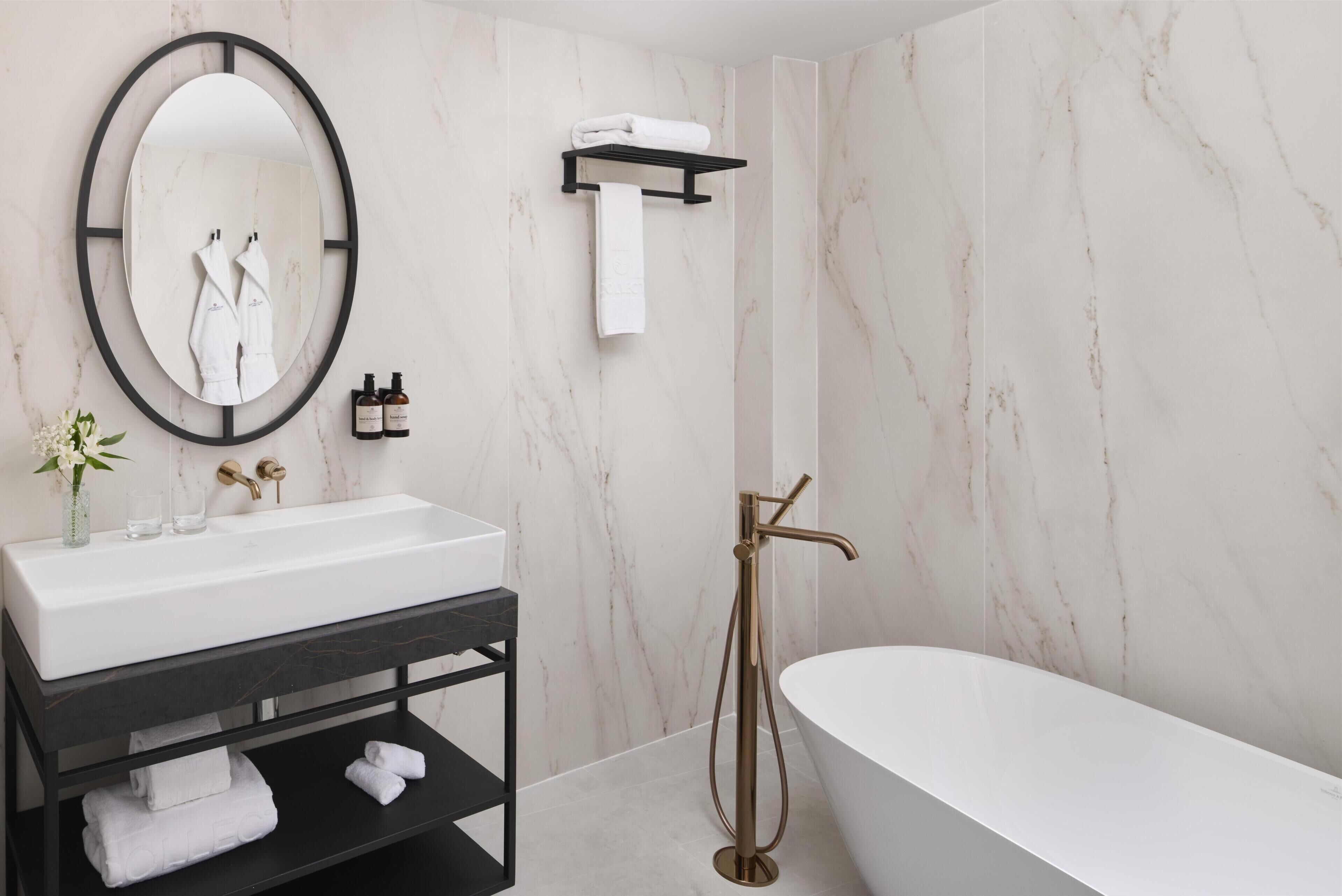 A modern bathroom with a freestanding bathtub, a large oval mirror, a black vanity with a white sink, and marble walls.