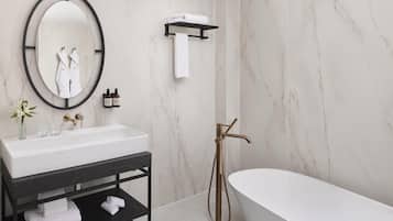 A modern bathroom with a freestanding bathtub, a large oval mirror, a black vanity with a white sink, and marble walls.
