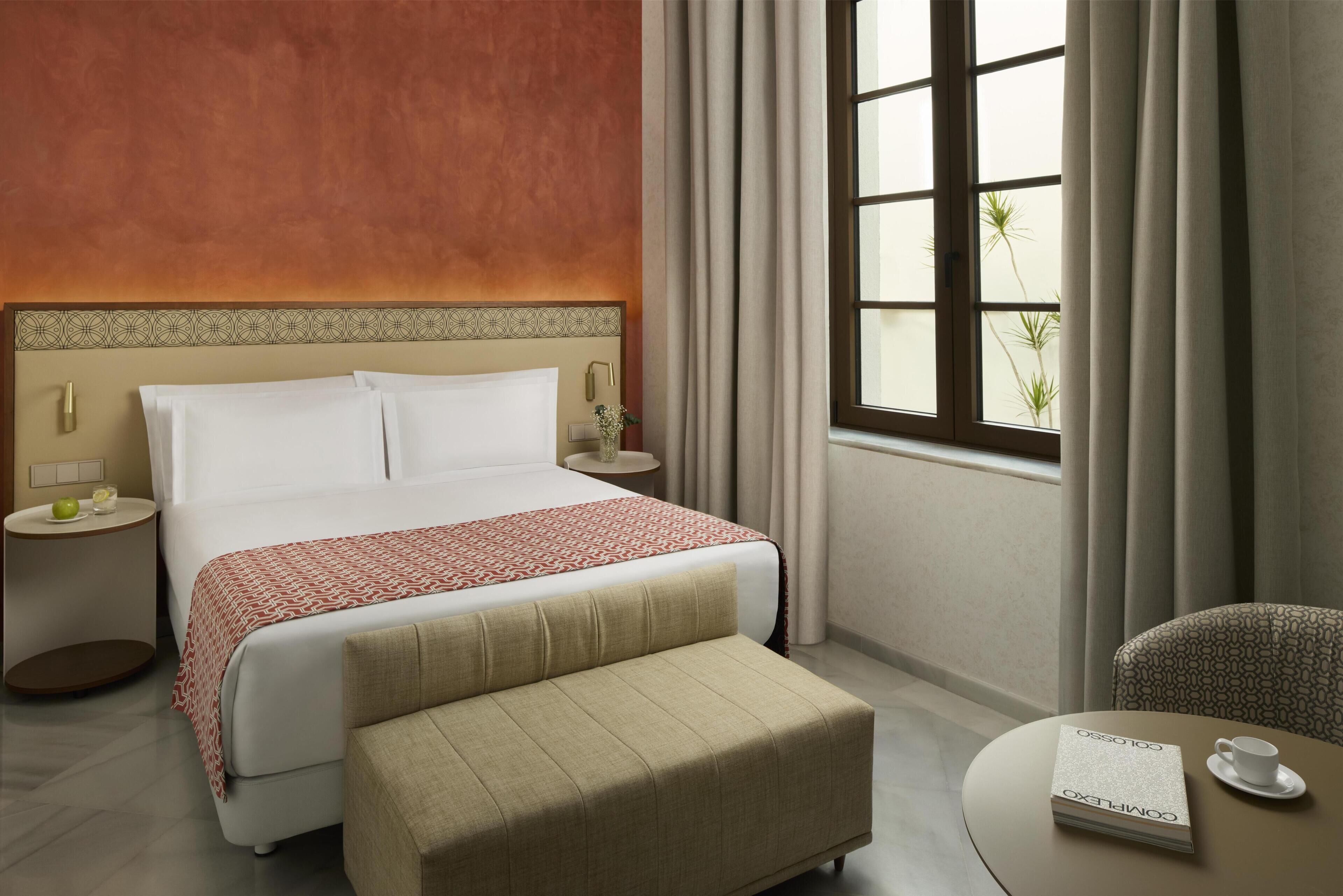 A hotel room with a bed, bedside tables, a sofa, a window with curtains, and a view of greenery outside.