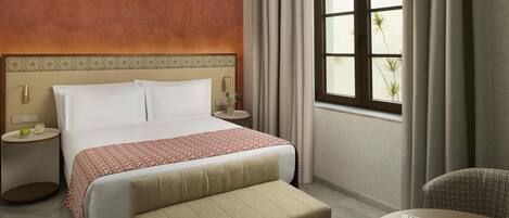 A hotel room with a bed, bedside tables, a sofa, a window with curtains, and a view of greenery outside.